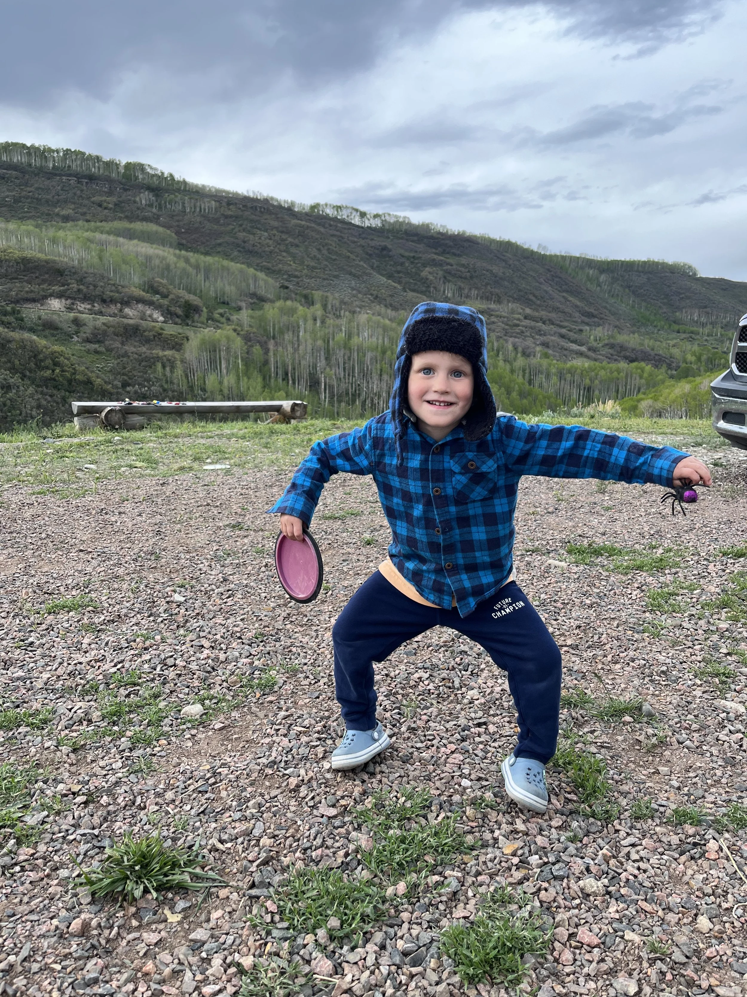A young boy in a blue plaid shirt and navy pants with the words "Future Champion" standing on a gravel and grass field, holding a frisbee and a small spider toy, with a scenic mountain landscape and cloudy sky in the background.
