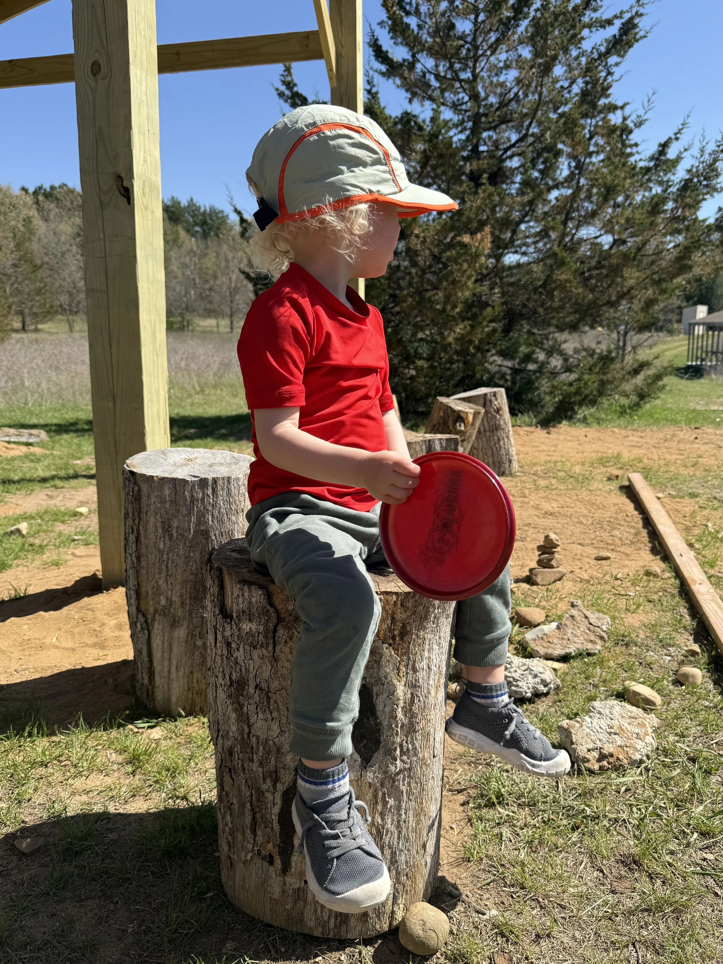 Young child with curly blonde hair wearing a white hat with orange trim, red shirt, gray sweatpants, and sneakers, sitting on a tree stump outdoors, holding a red frisbee, with a background of trees and blue sky.