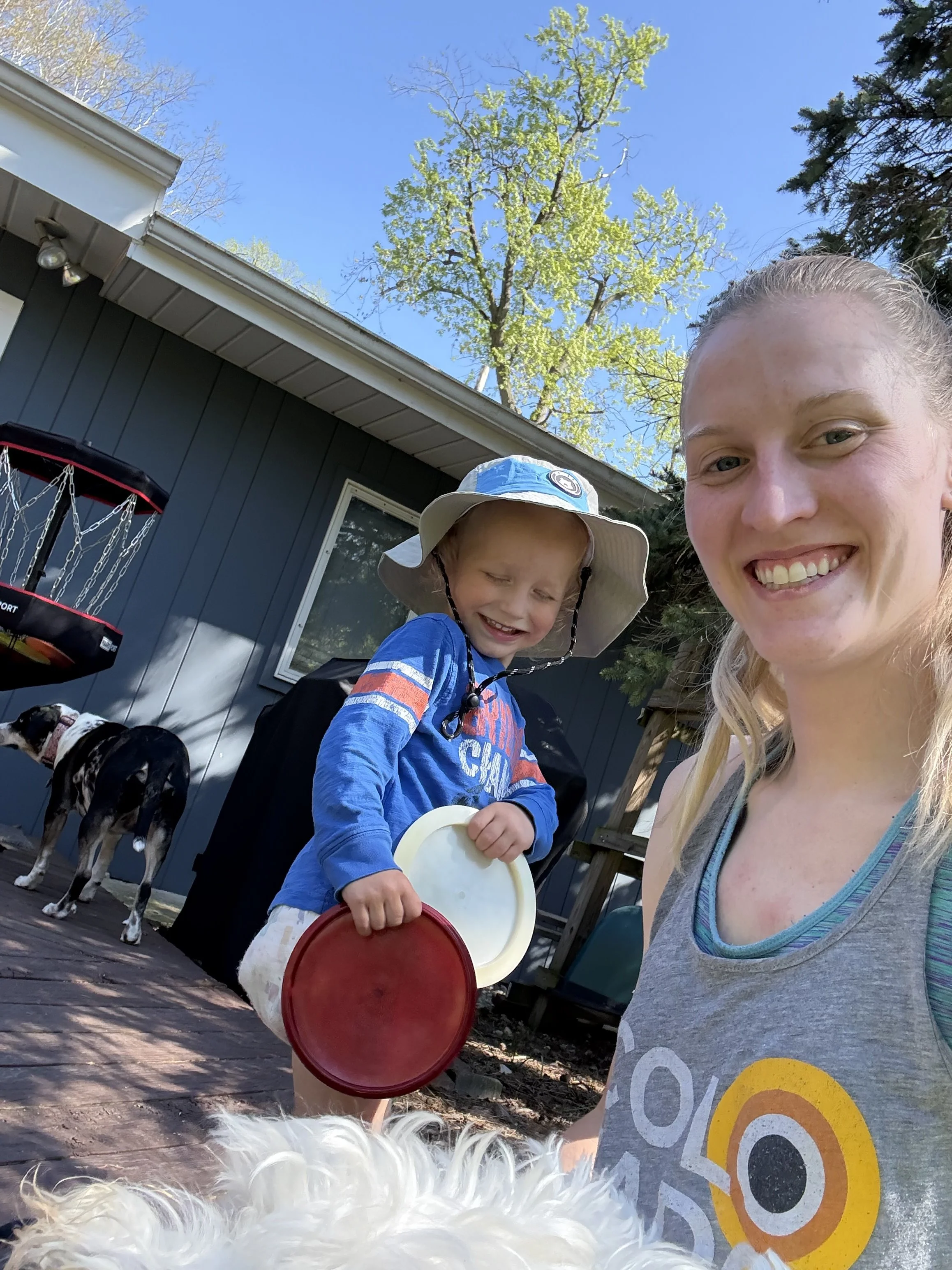 A smiling young woman and a boy with a sun hat are outdoors on a sunny day, with trees and a house in the background. The boy is holding colored lids and a container, and a dog is partially visible in the foreground.
