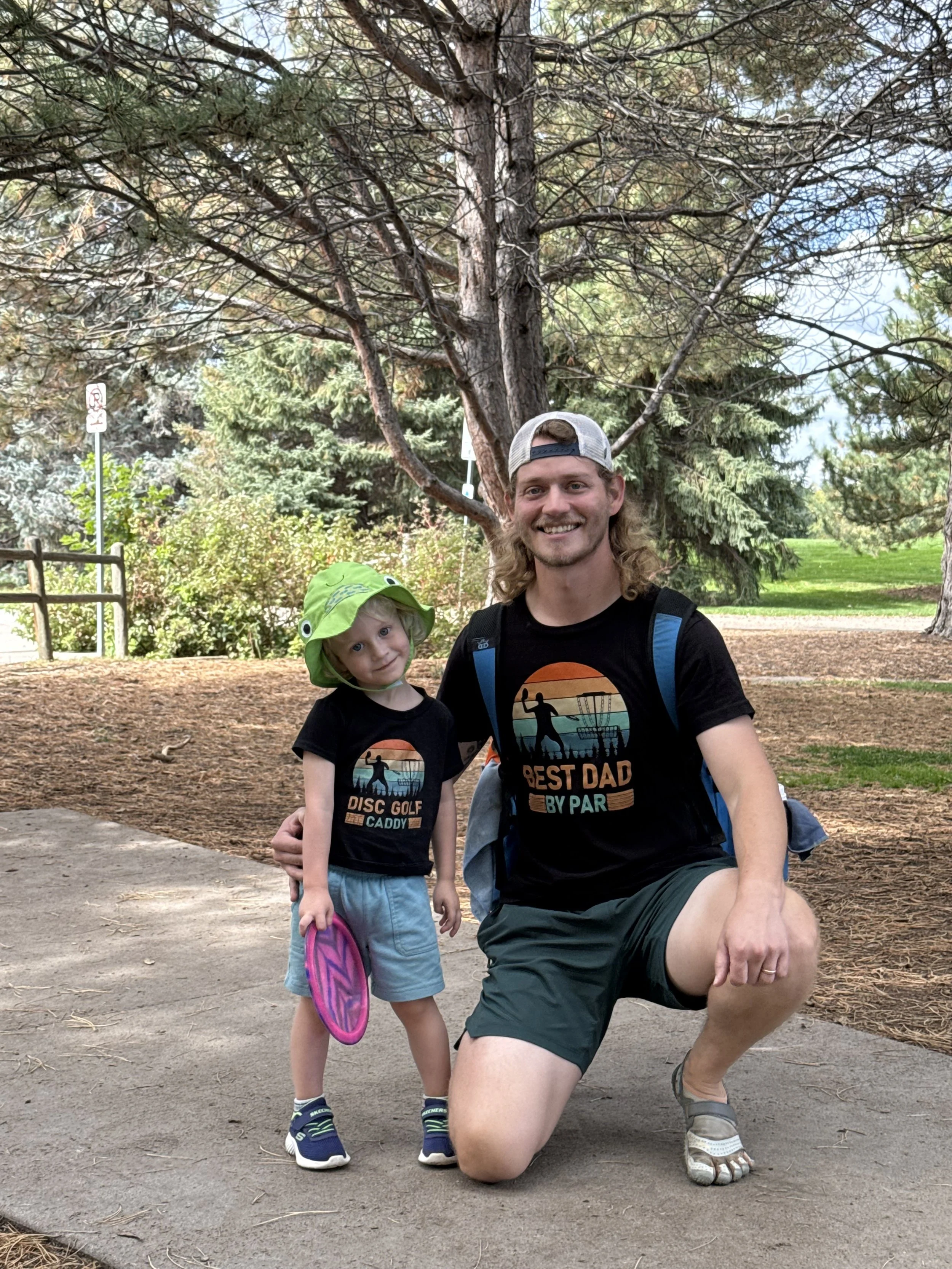 A man and a young girl posing outdoors. The man is kneeling on one knee, smiling at the camera, wearing a black t-shirt with a disc golf graphic and text, and a backwards baseball cap. The girl is standing next to him, also smiling, wearing a black t-shirt with a disc golf graphic, light-colored shorts, sneakers, and a bright green hat with eyes. They are in a wooded park with trees and a dirt path.