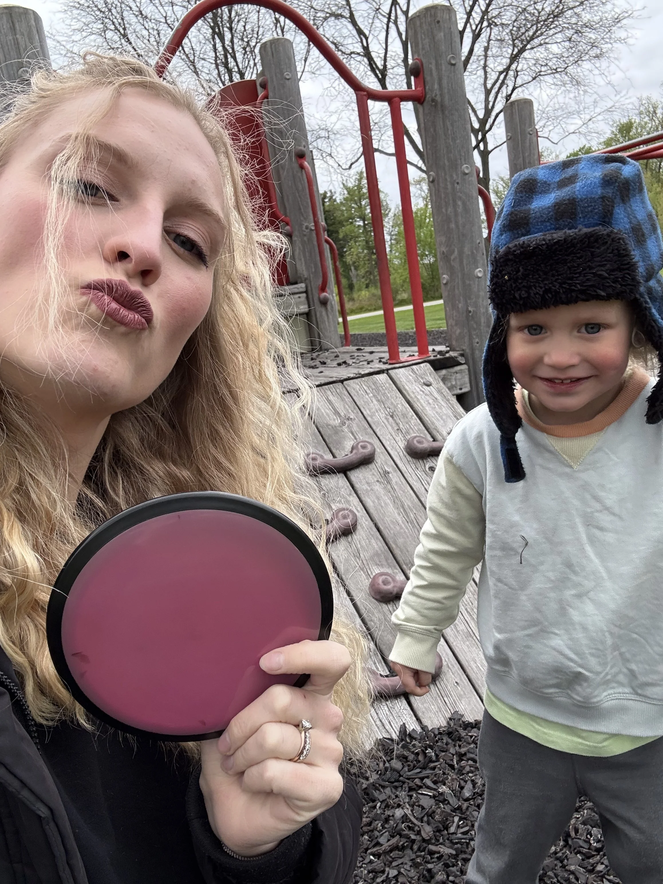 A woman with blonde curly hair and purple lipstick holding a pink reflective object, and a young boy with a blue and black checkered winter hat, holding hands outside at a playground with trees and overcast sky in the background.