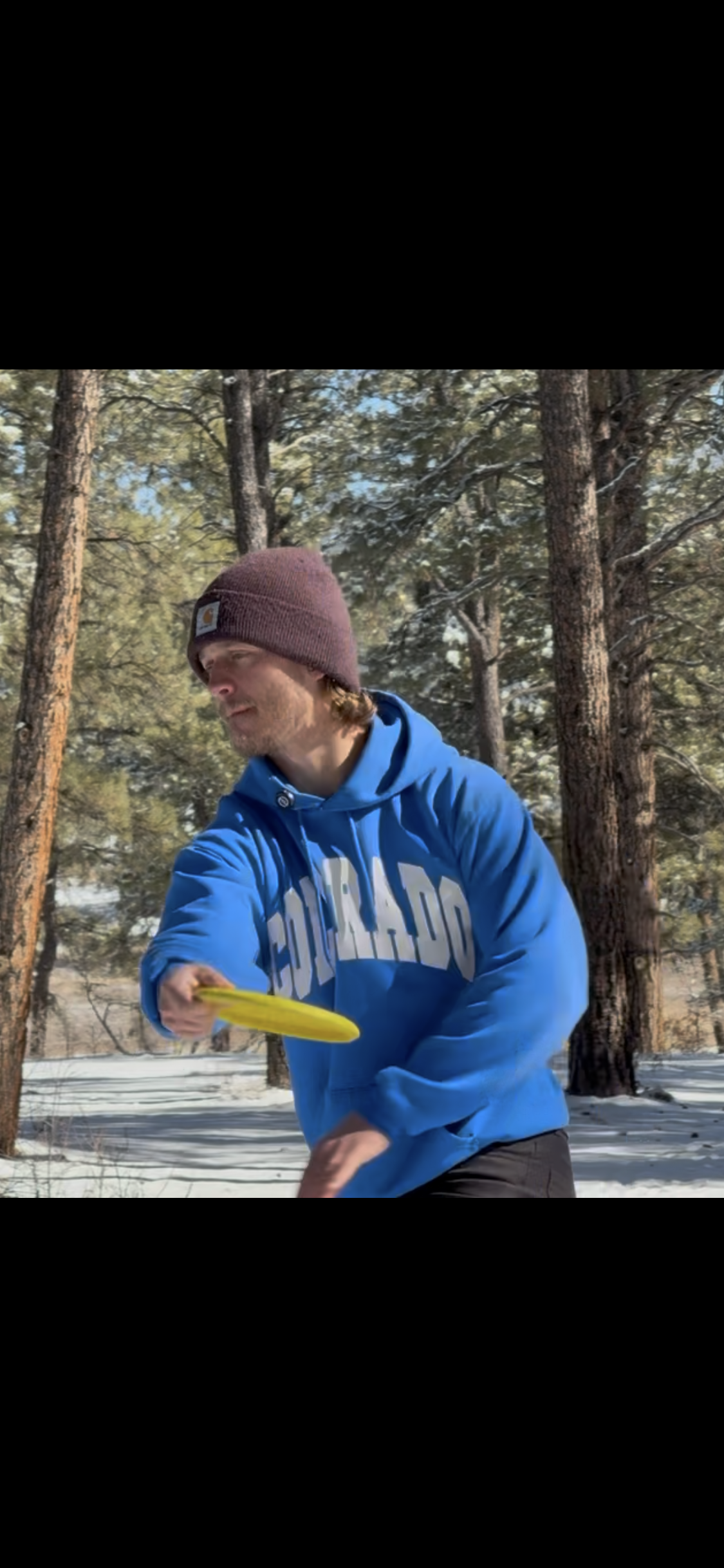 A young man in a blue hoodie and maroon knit cap throwing a frisbee in a snowy forest.