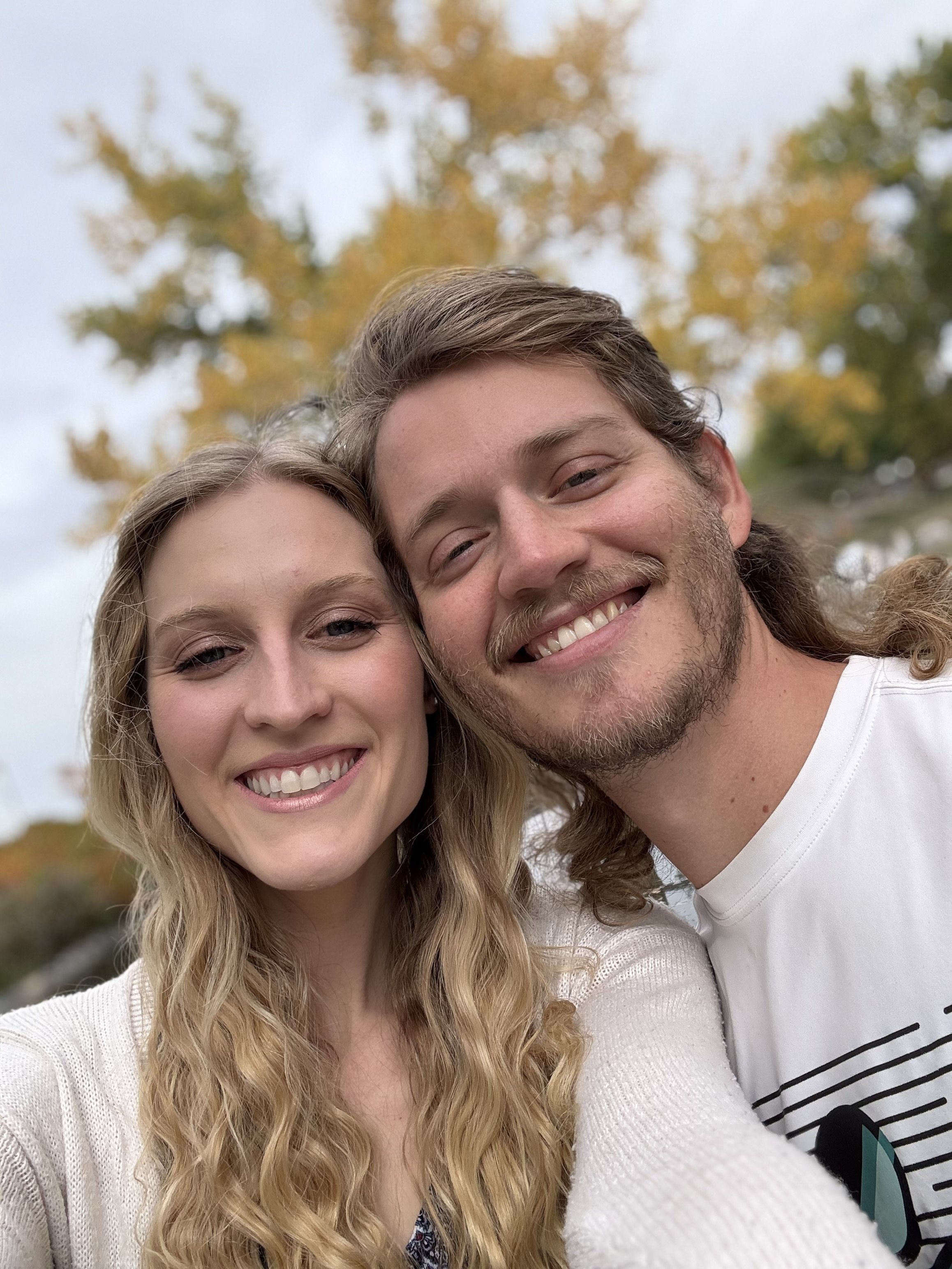 A young woman and a young man taking a selfie outdoors on a cloudy day with trees showing fall colors in the background.