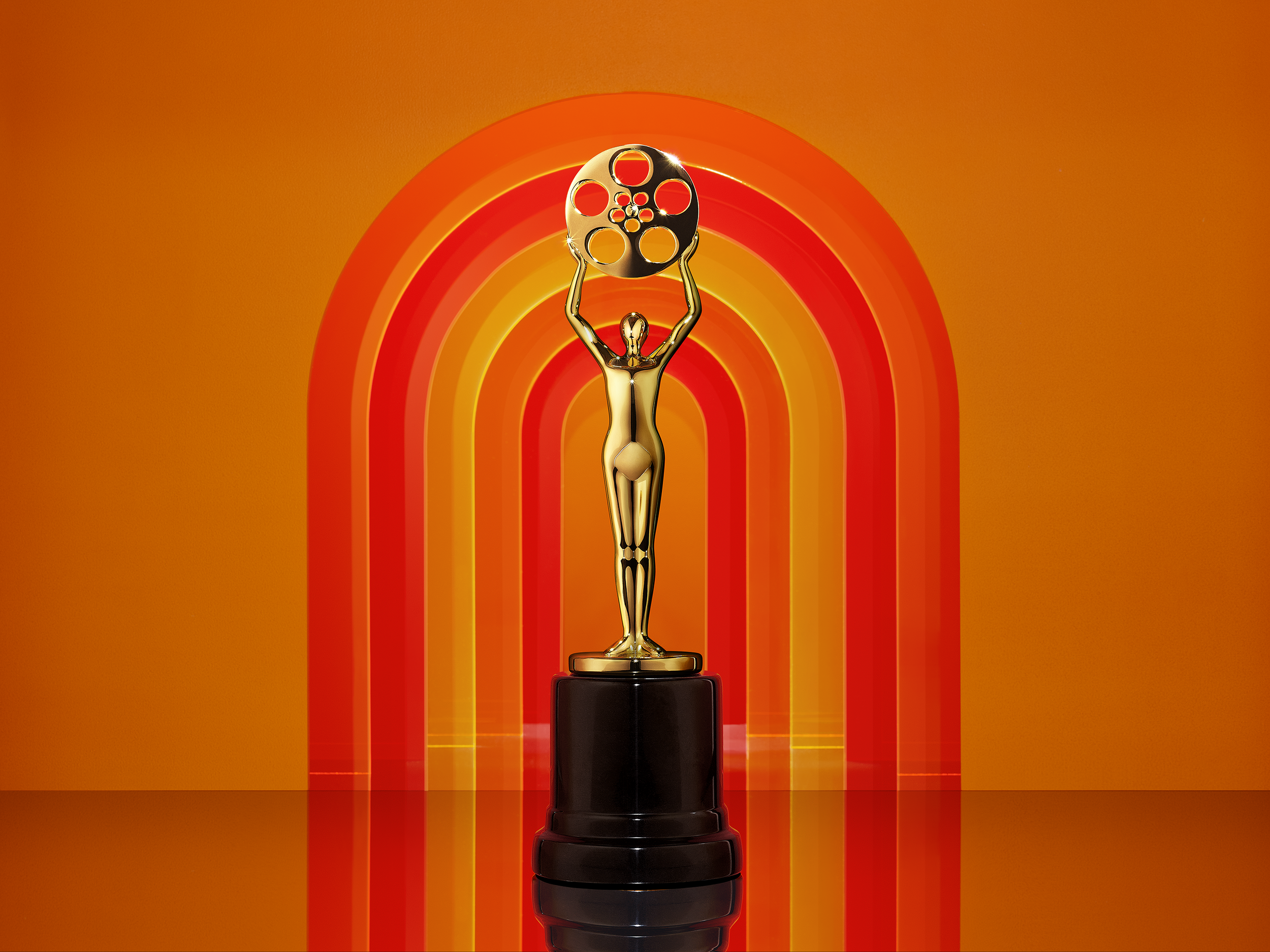 A shiny, golden humanoid figure holding a film reel above its head stands on a black pedestal against a warm orange background with concentric arches.
