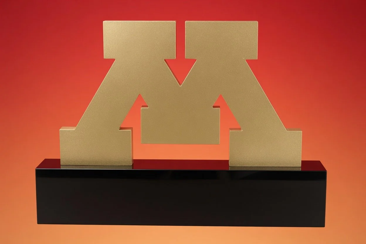 A sculpture award of the letter 'M' with arrows pointing inward on a black base, set against a red-orange background.