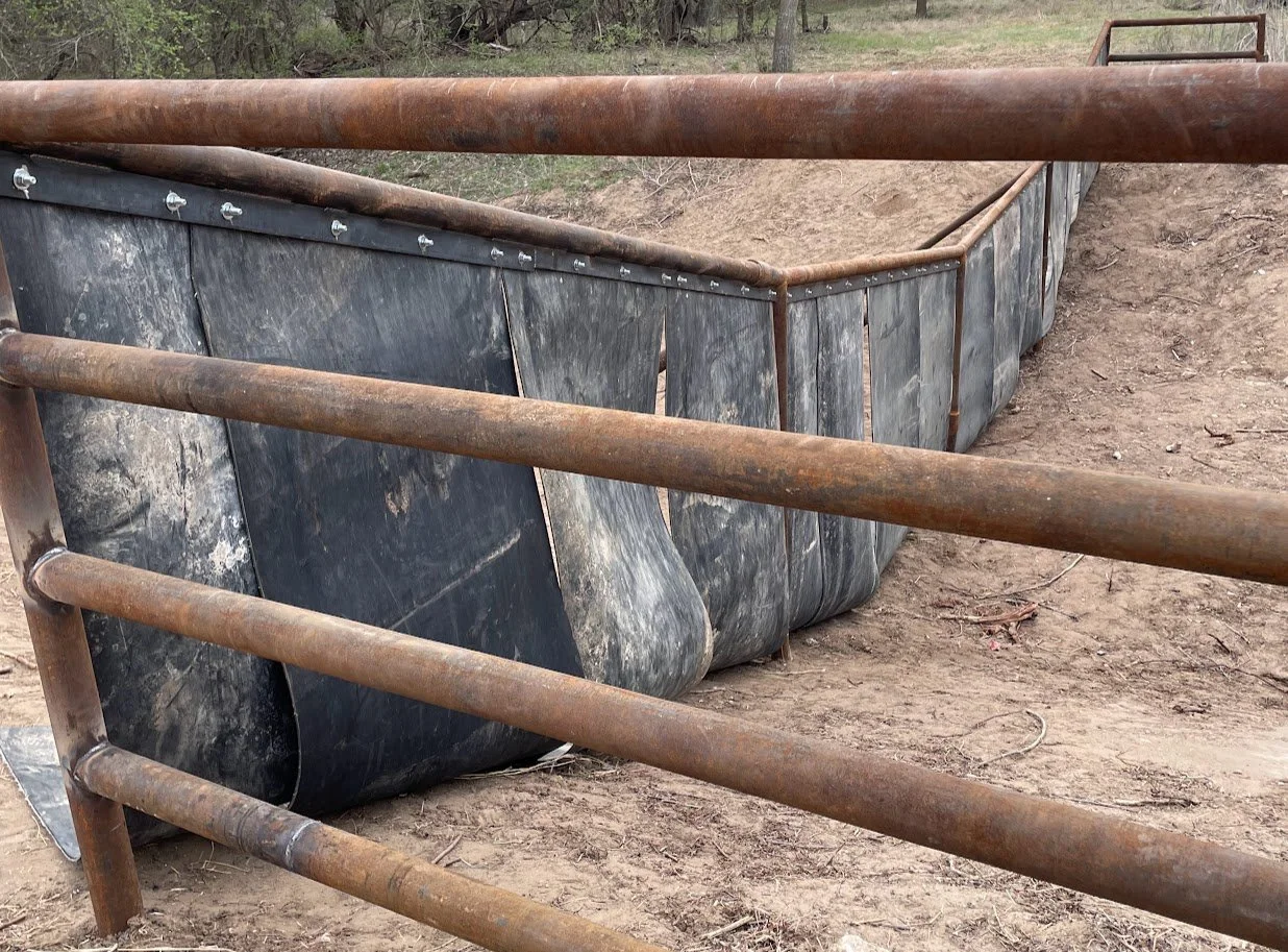 A dirt rode with a metal fence along the side. The fence has three horizontal bars and vertical supports and surrounds a rectangular dug-out area lined with black rubber or plastic sheeting.