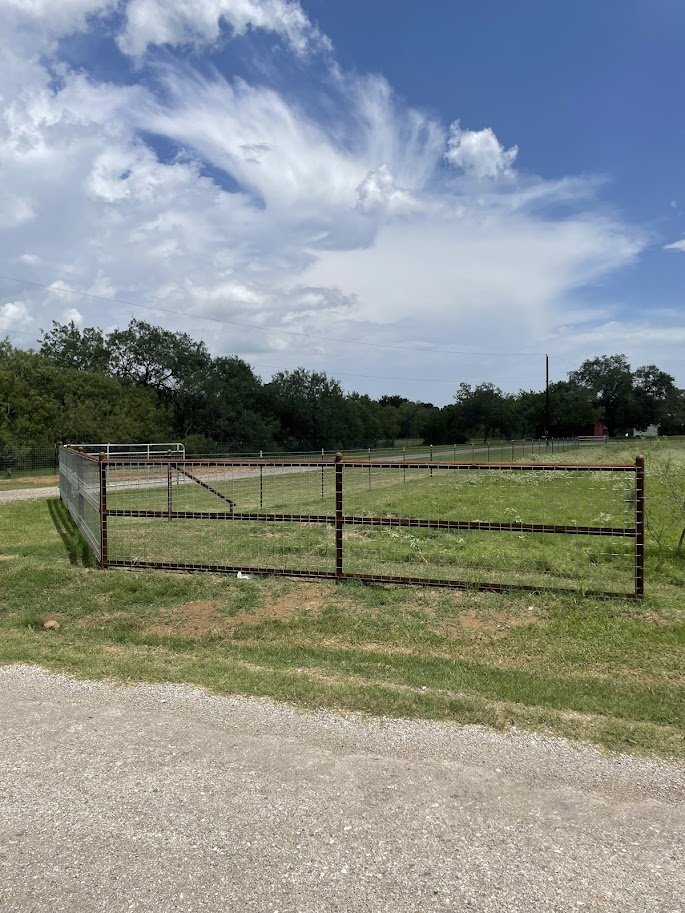 A fenced area on a grassy field with a dirt road in the foreground and trees in the background, under partly cloudy sky.
