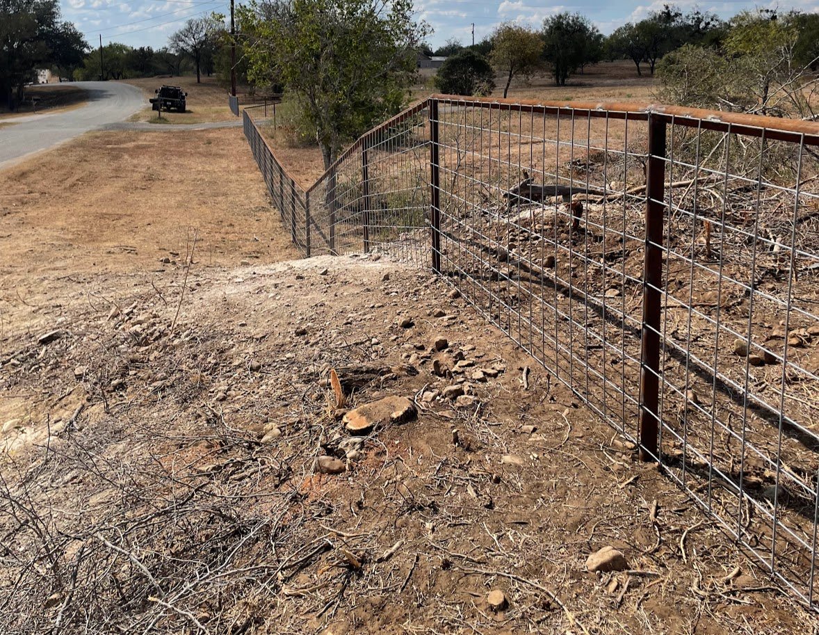 A metal wire fence along a dirt path on a hillside, with a road and a vehicle in the background, surrounded by dry grass and sparse trees.