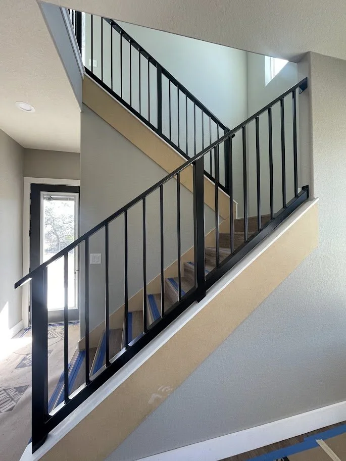 Interior view of a staircase with black metal railing, light-colored wooden steps, and a wall with a window letting in natural light.
