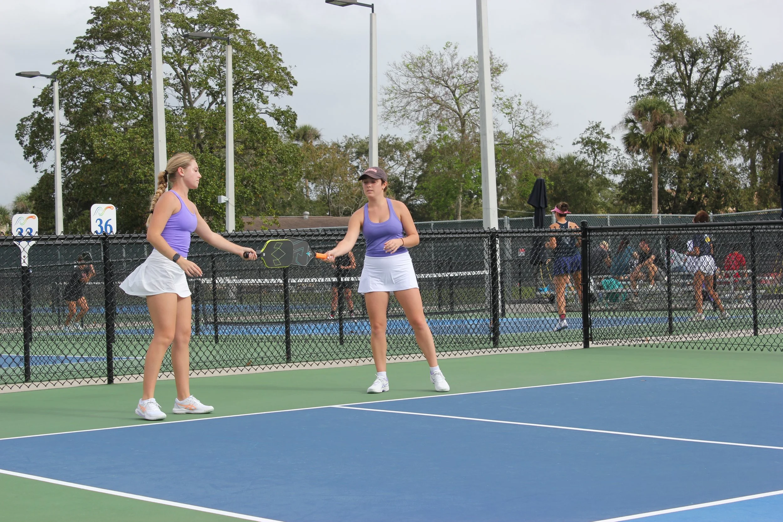 Two young women wearing purple tank tops and white skirts playing pickleball on an outdoor court, with a chain-link fence and trees in the background.