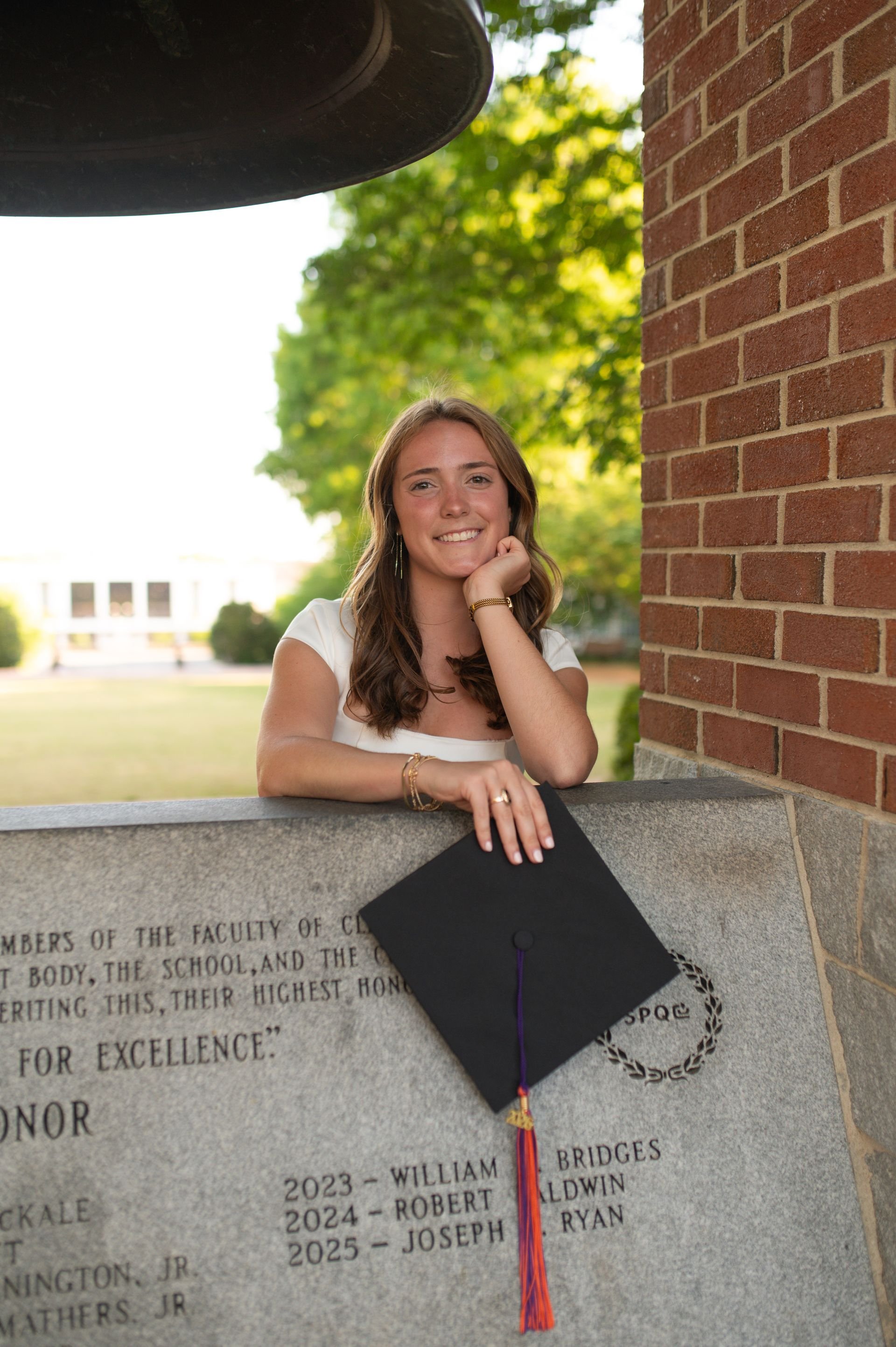 A young woman with brown hair, wearing a white shirt, inside., sitting at a stone monument or plaque with an engraved list of names and years, with a brick wall to the right and green trees in the background, holding a graduation cap with a tassel.