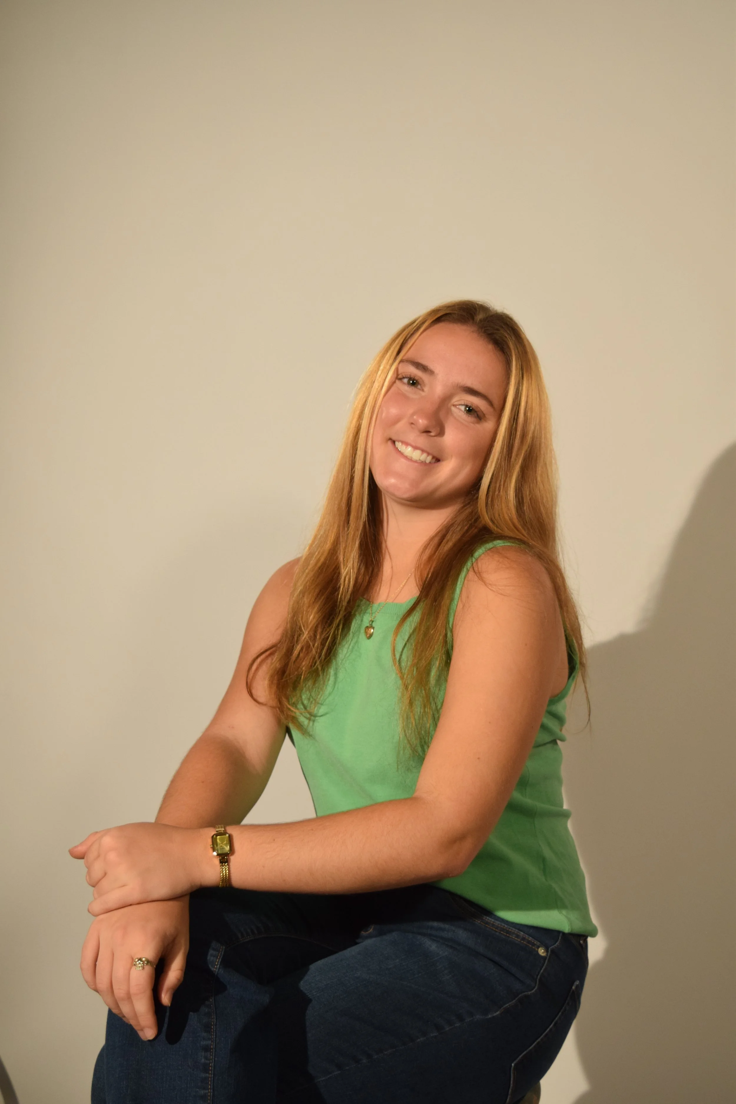 A young woman with long red hair, wearing a green sleeveless top and dark jeans, smiling and sitting against a plain beige wall.