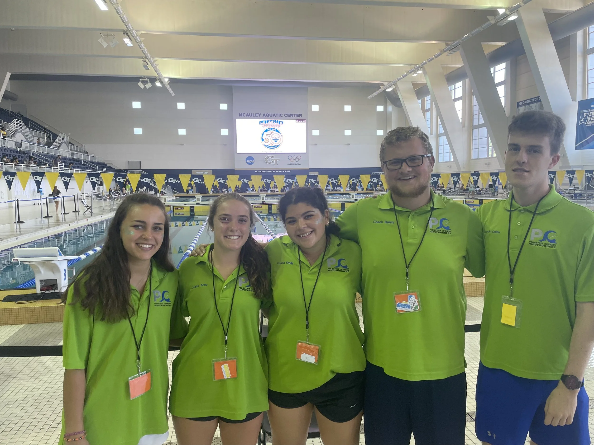 A group of five young women and one young man standing together inside an indoor swimming pool facility. They are wearing bright green shirts with blue logos and are smiling at the camera. The background shows swimming lanes, a large electronic score
