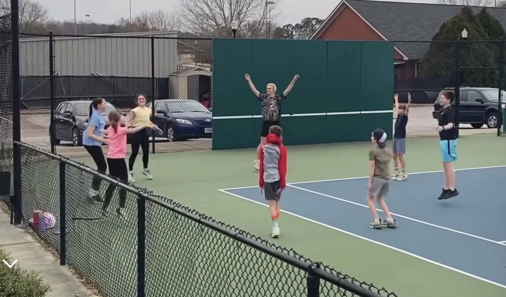 Group of children and an adult playing tennis on an outdoor court during daytime, surrounded by a black chain-link fence, with parked cars and residential buildings in the background.