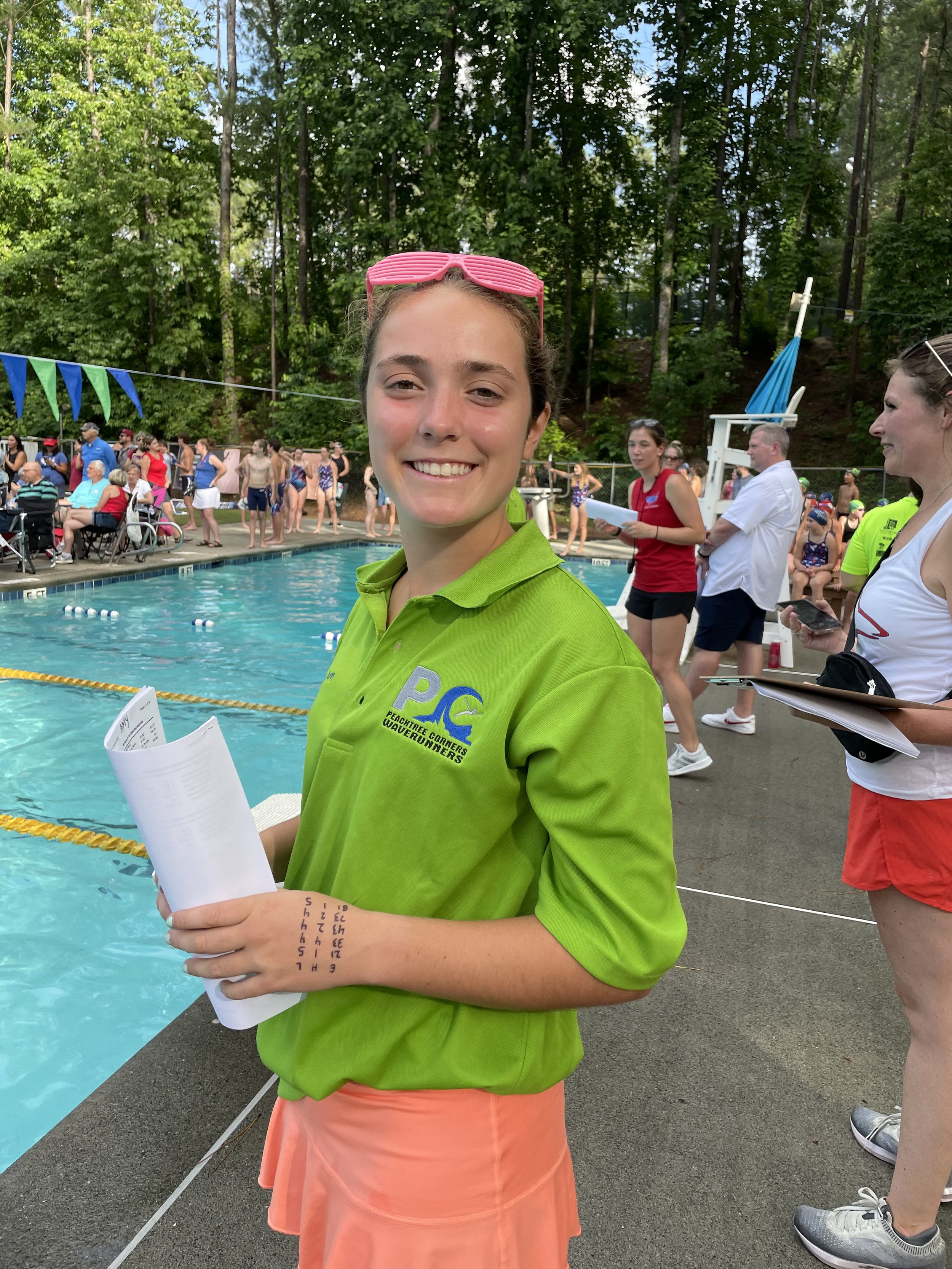 A young woman with a pink sunglasses on her head, wearing a bright green shirt, and holding a paper, standing poolside at a swimming event. In the background, people in swimwear and officials are gathered near the pool, with trees surrounding the are