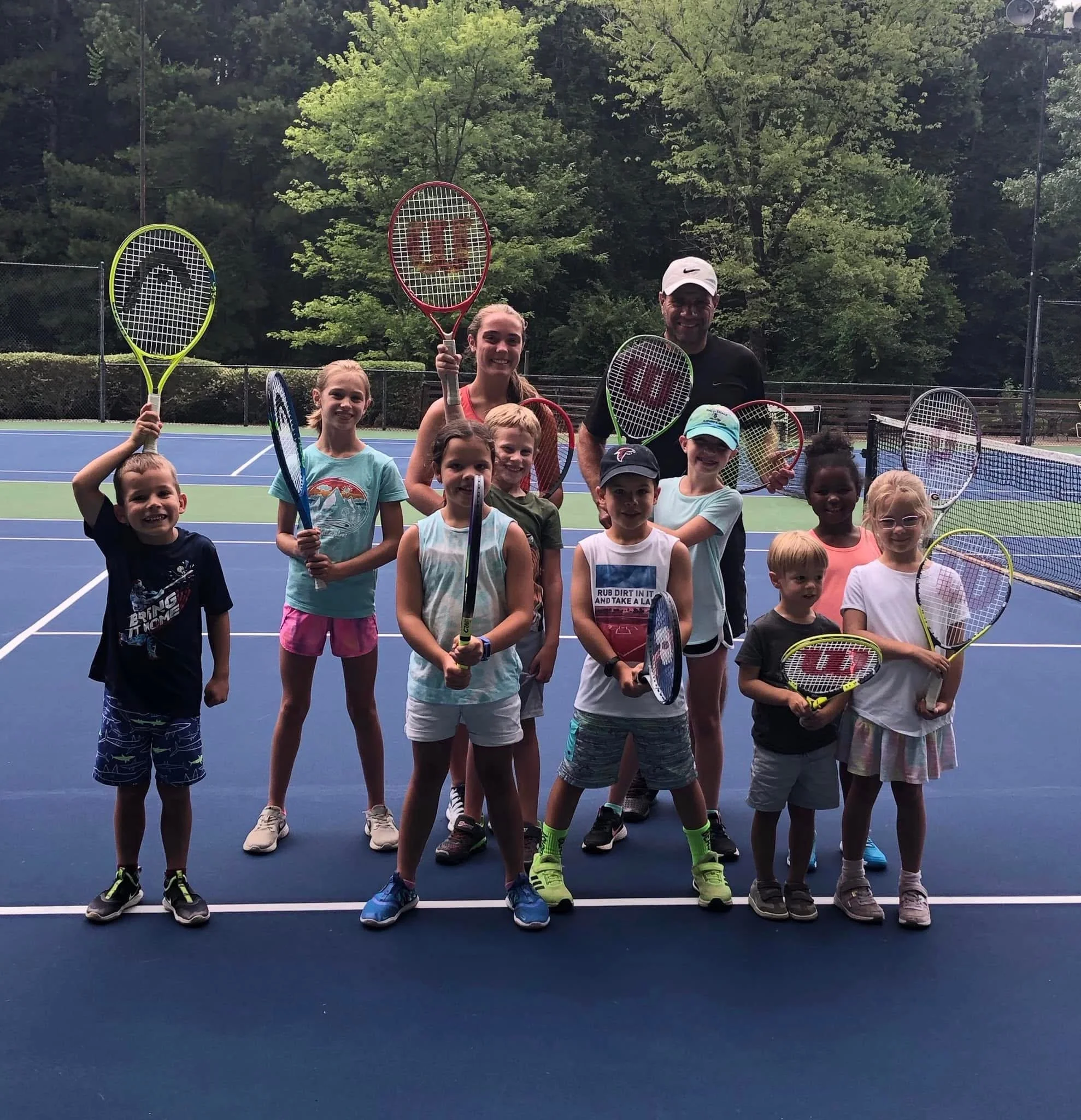 Group of children and a coach posing on a tennis court, holding tennis rackets, with trees in the background.