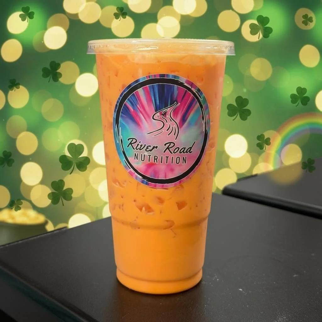 An orange blended drink in a clear plastic cup with a logo reading 'River Road Nutrition'. The background has a green theme with shamrock shapes and bokeh lights, suggesting a festive or St. Patrick's Day setting.