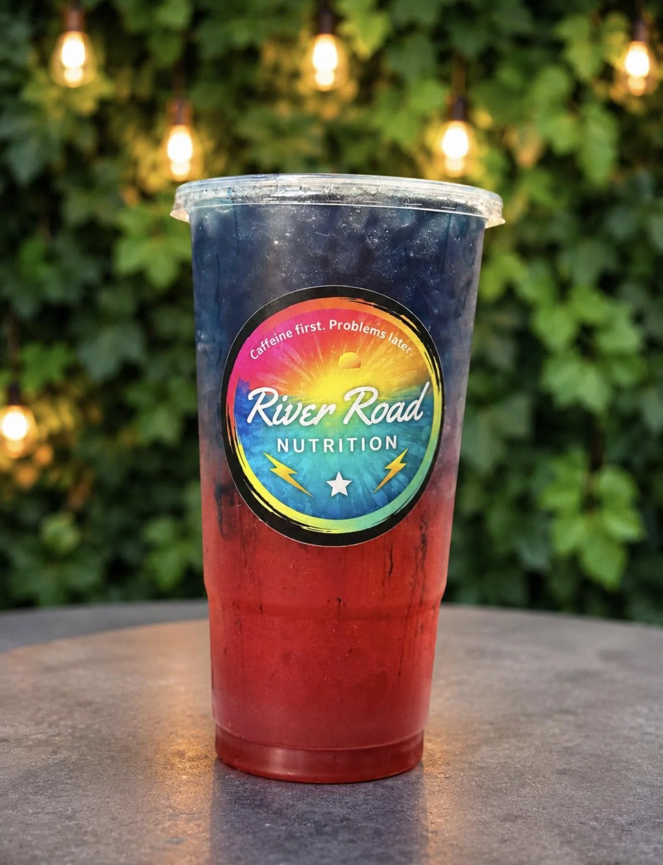 A colorful iced drink in a plastic cup with a logo that reads 'River Road Nutrition' and the tagline 'Caffeine first. Problems later.' The background features green foliage and string lights.
