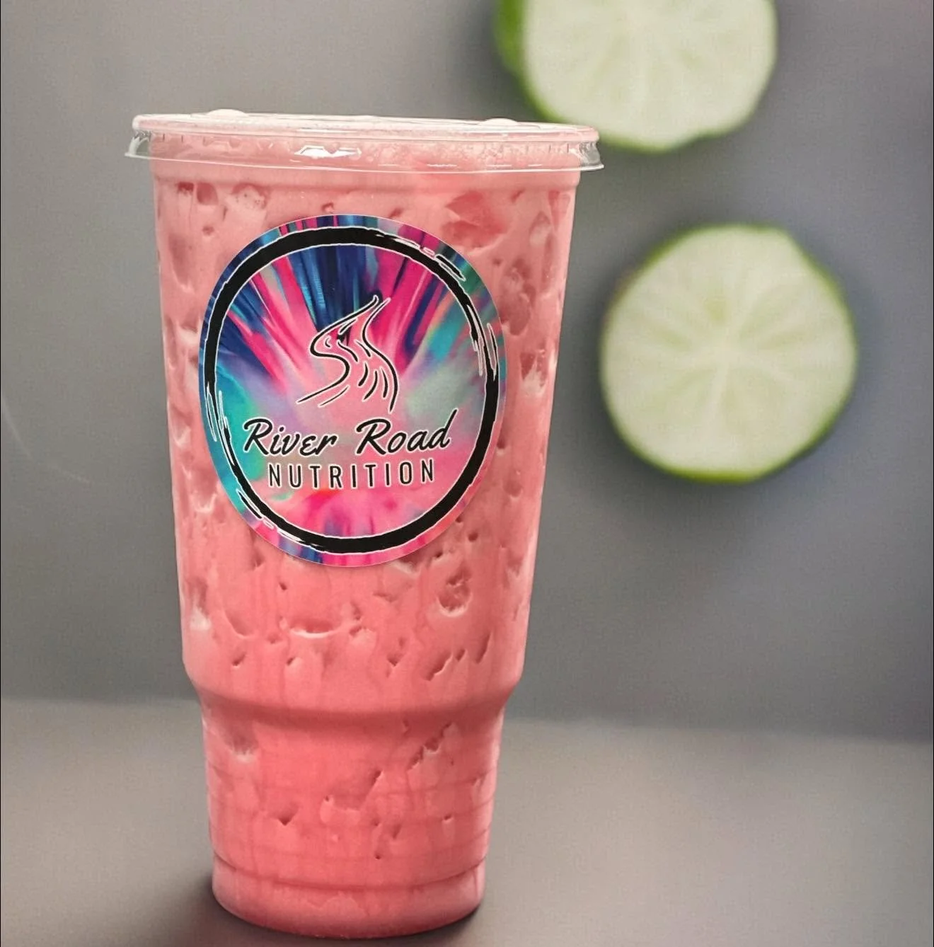 A pink smoothie in a clear cup with a colorful logo of River Road Nutrition, with two lime slices in the background on a gray surface.