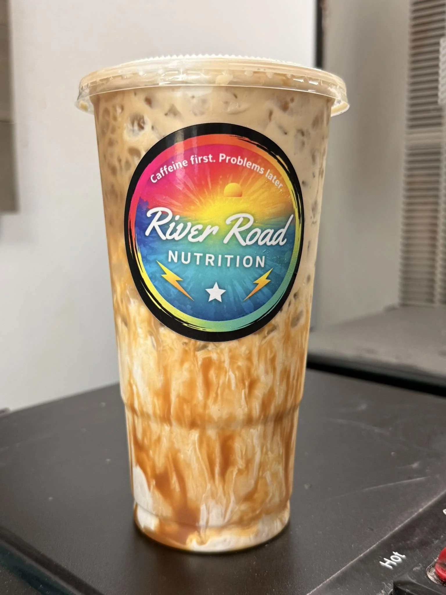 A clear plastic cup filled with a caramel-colored iced beverage, placed on a black surface near a microwave.