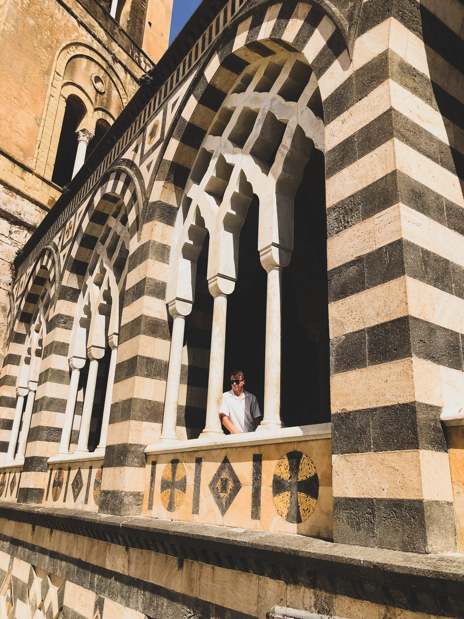 A man wearing sunglasses and a white shirt standing on a decorative balcony with black and white striped arches and stone columns, part of an ornate historic building with intricate window details.