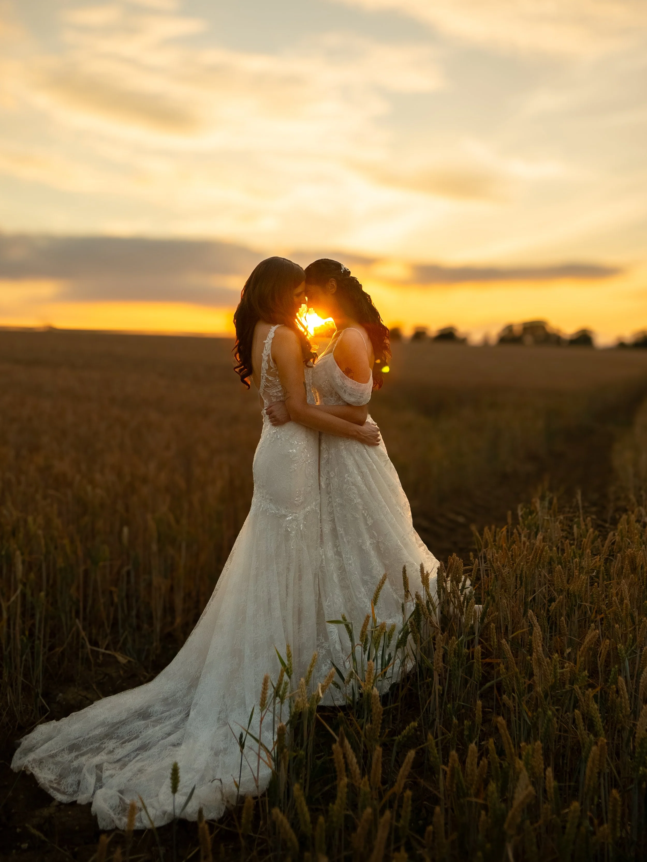 Two women wearing white wedding dresses holding each other in a field during sunset.