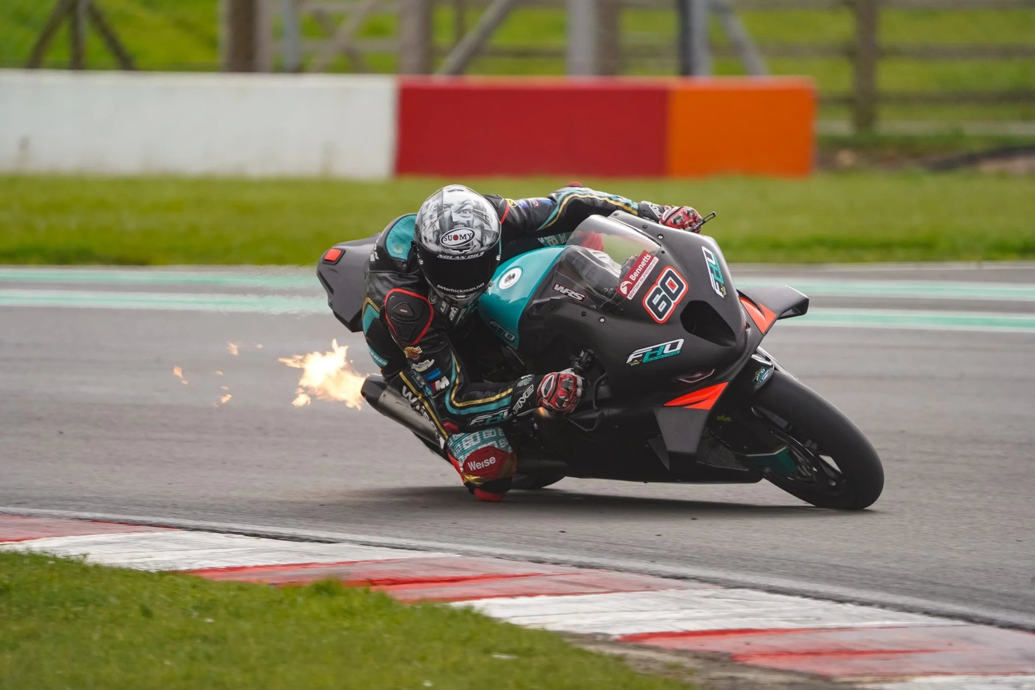 Motorcycle racer leaning into a turn on a race track, with sparks flying from the bike's exhaust, background showing green grass and a red and white wall.