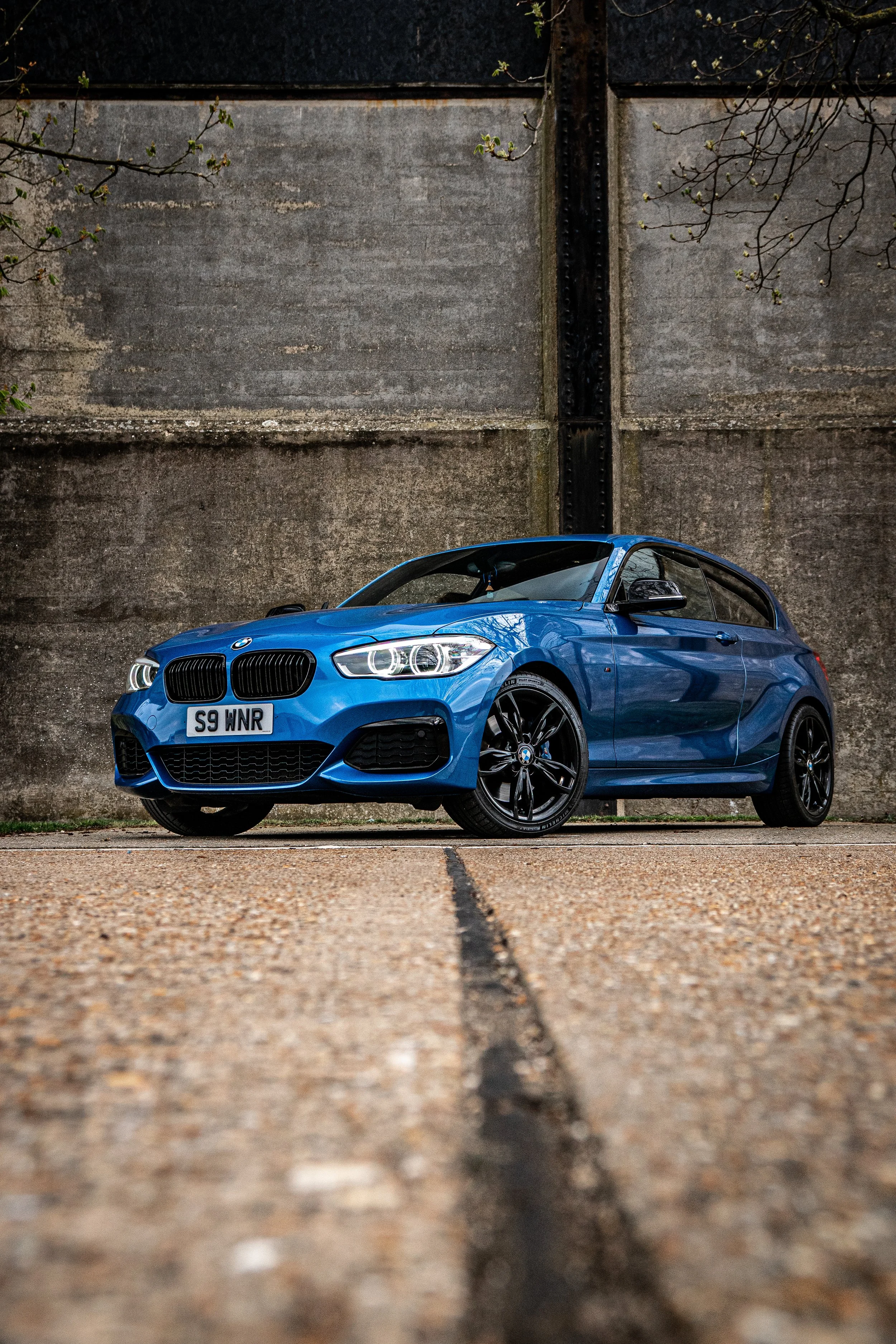 Blue BMW car parked on concrete pavement in front of a large concrete wall with some tree branches visible at the top.
