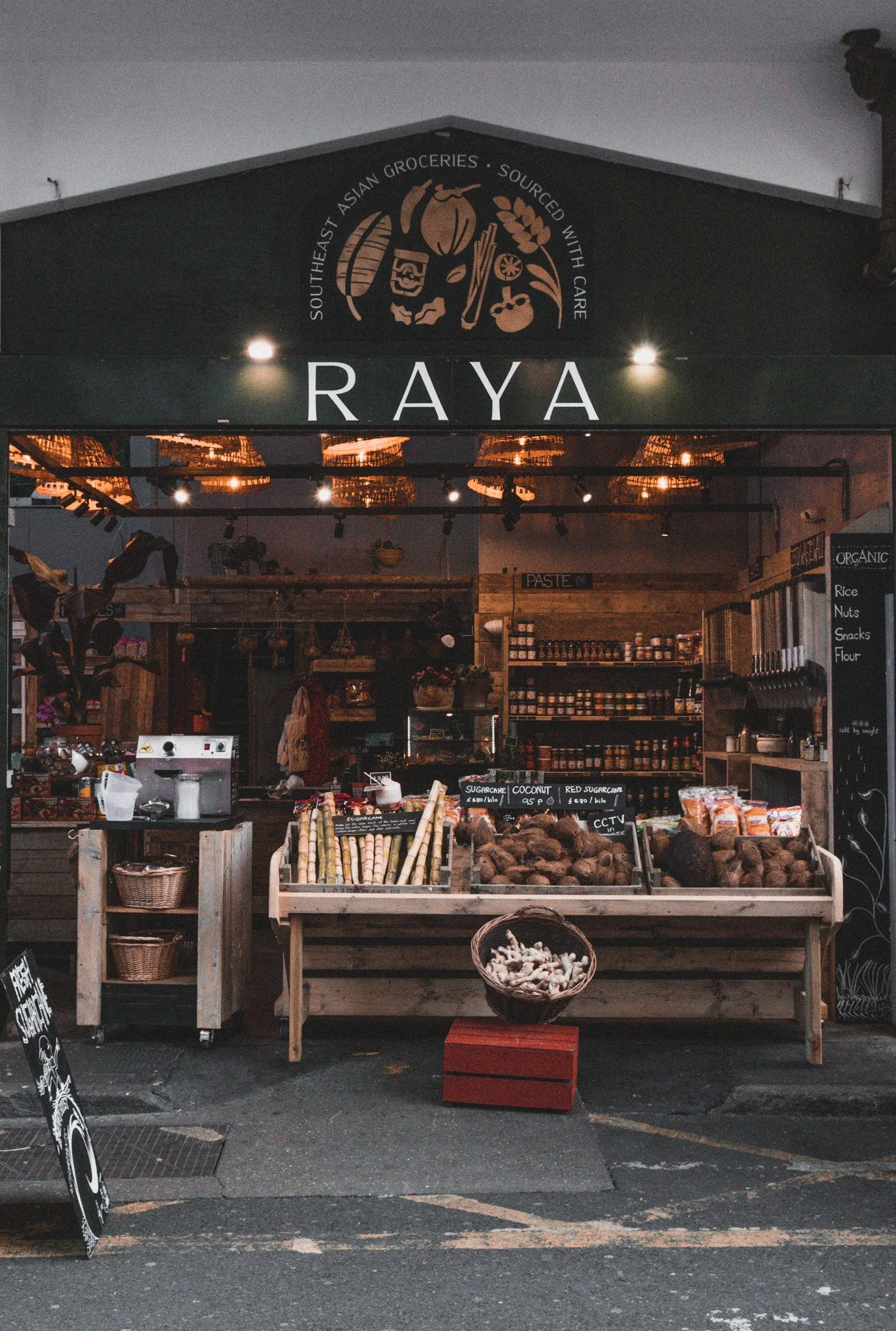 A street vendor stall with various fresh produce including roots, ginger, and other vegetables on display. The stall has a sign with the name 'RAYA' and features a black awning with a logo above. The interior is warmly lit with hanging lights, and there are shelves with jars and containers behind the stall.