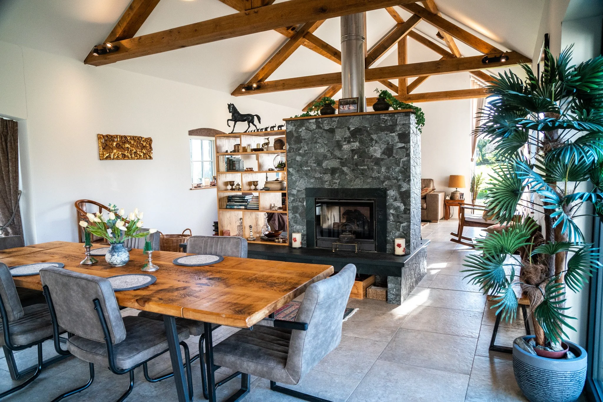 Living room with a wooden dining table with gray chairs, a stone fireplace, tall houseplants, and decorative items on shelves and around the room. Exposed wooden beams on the ceiling.