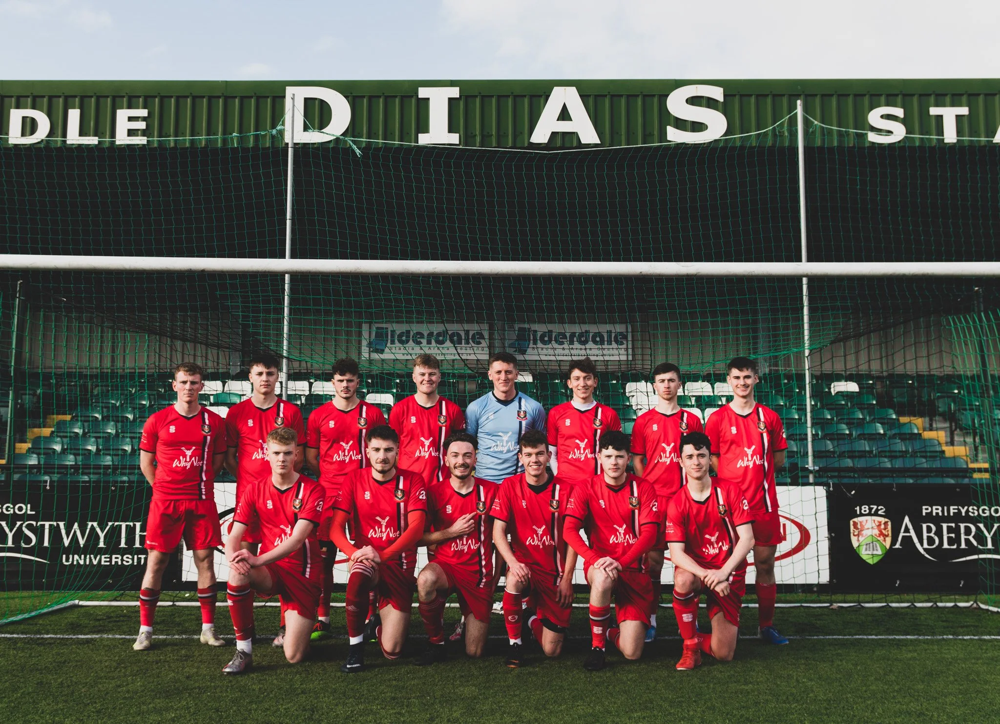 A group of young male soccer players in red uniforms pose for a team photo on a soccer field, with a green fence and stadium seating in the background.