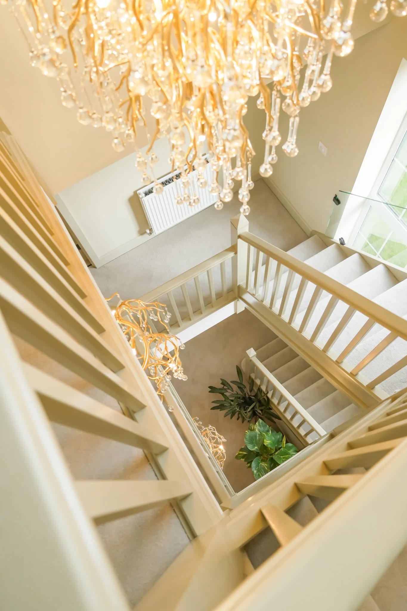 View looking down from the top of a staircase showing a chandelier hanging from the ceiling, a radiator below, stairs leading down, and a large window with a view outside. There are also potted plants in the area.