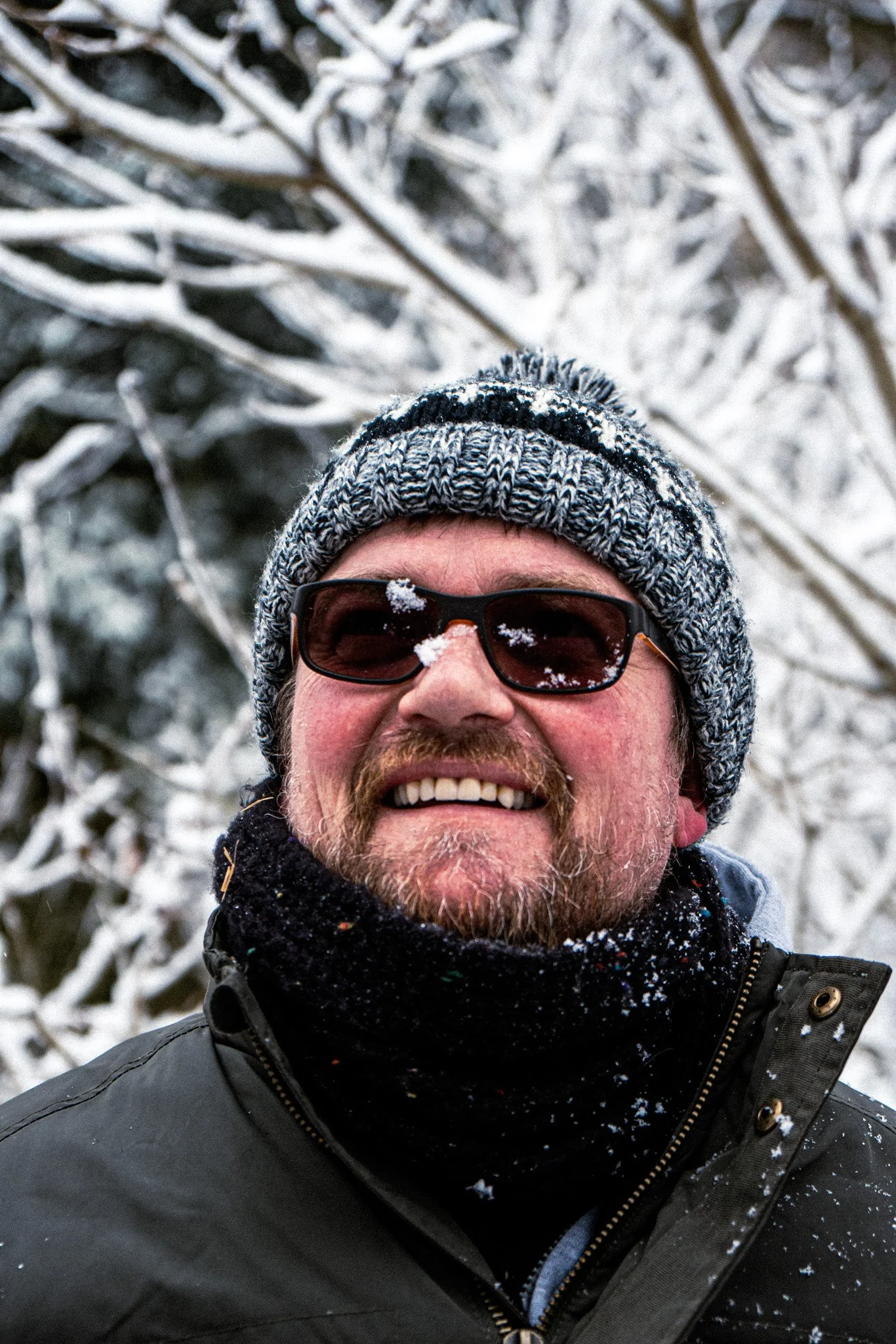 Close-up of a smiling man wearing a gray knit hat, sunglasses, and a black winter jacket, with snow on his face and clothing, standing outdoors with snow-covered trees in the background.