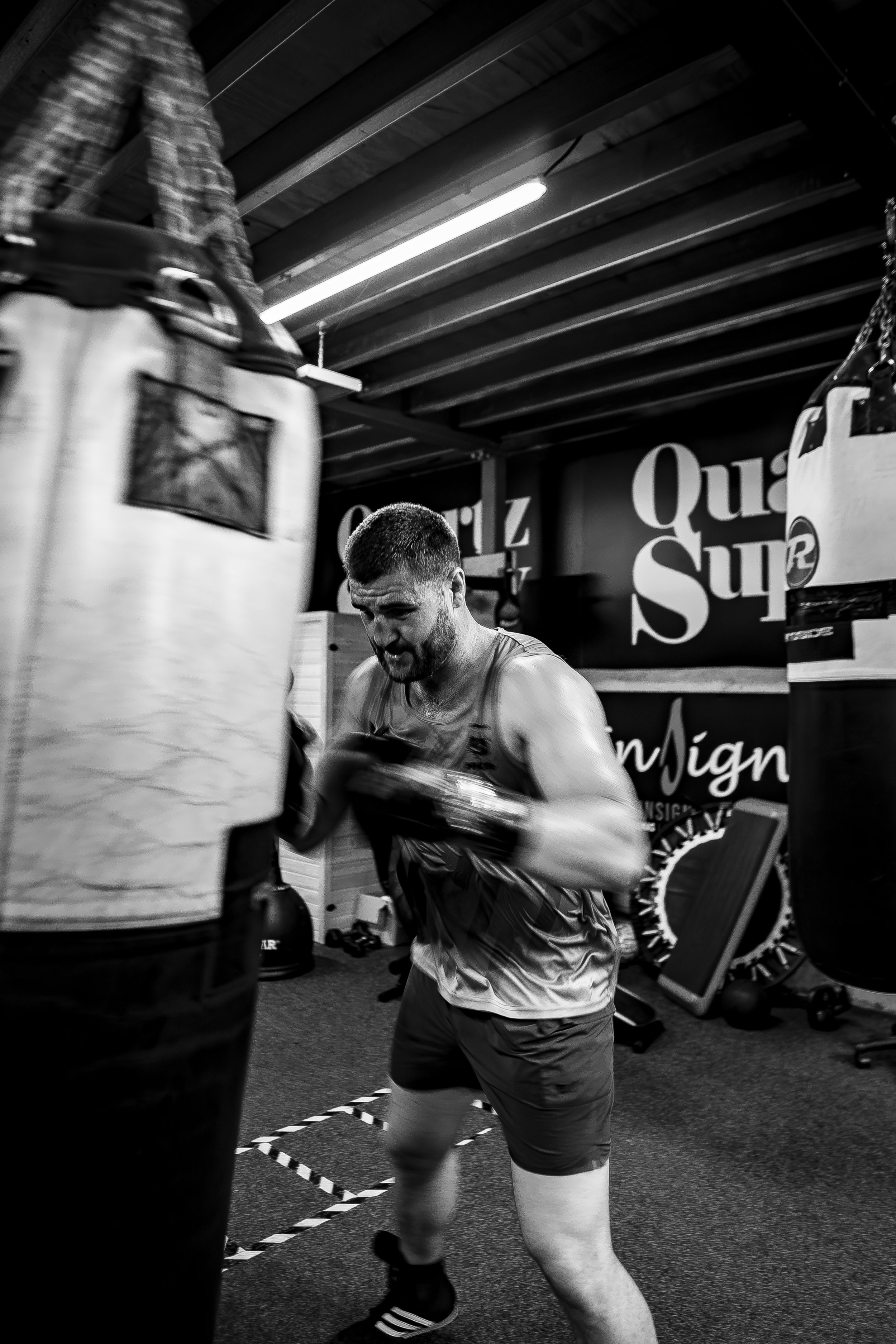 A man with a beard and short hair training in a gym, wearing a sleeveless shirt and shorts, aiming punches at a boxing heavy bag. The gym has motivational signs and equipment in the background.
