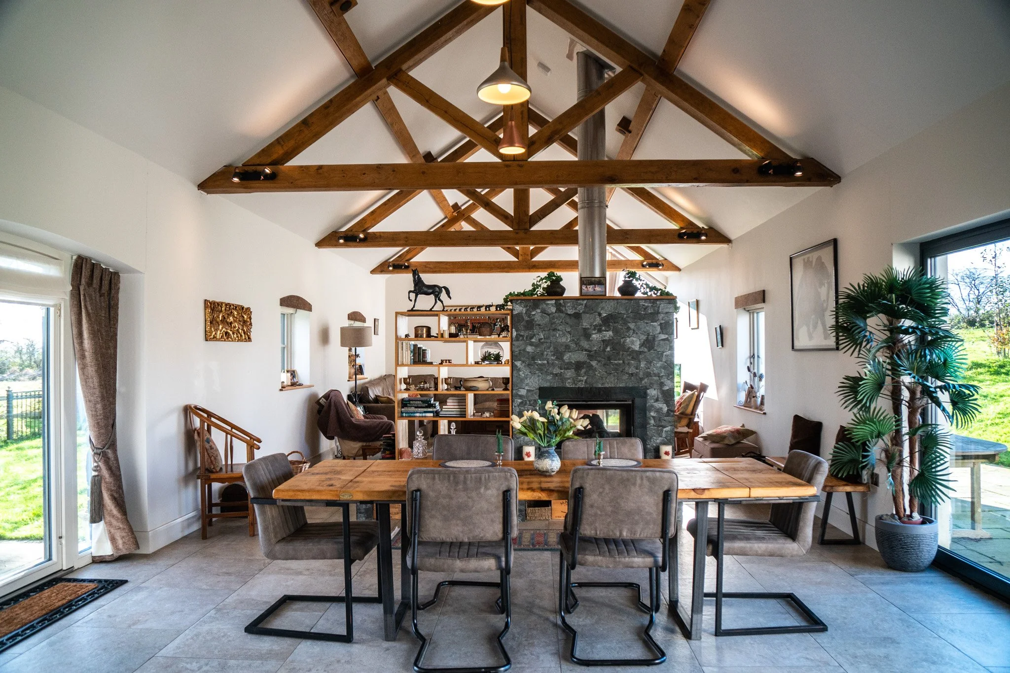 Interior of a modern living room with a high vaulted ceiling featuring wooden beams. A large stone fireplace is at the center with decorative items on top. The room has windows on both sides, allowing natural light, with curtains on the left window. A wooden dining table with six upholstered chairs is in the foreground, decorated with a flower centerpiece and candles. There's a plant on the right and various decorative items on shelves and walls.