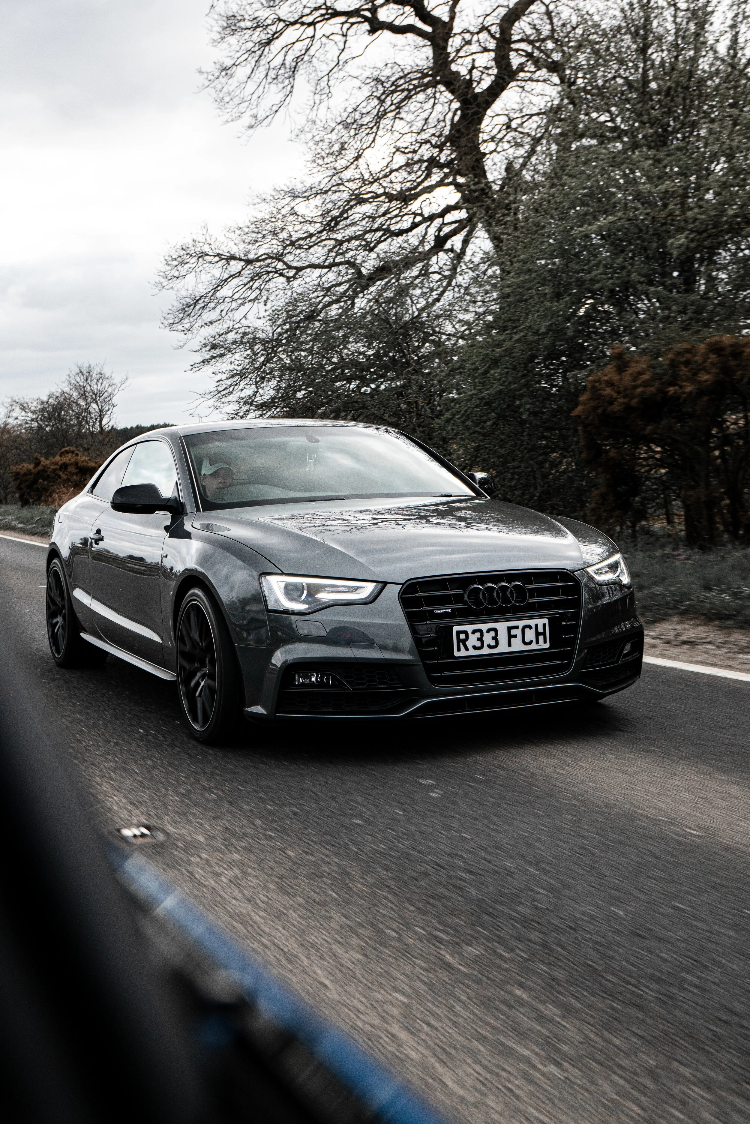 A gray Audi car driving on a highway with trees and cloudy sky in the background.