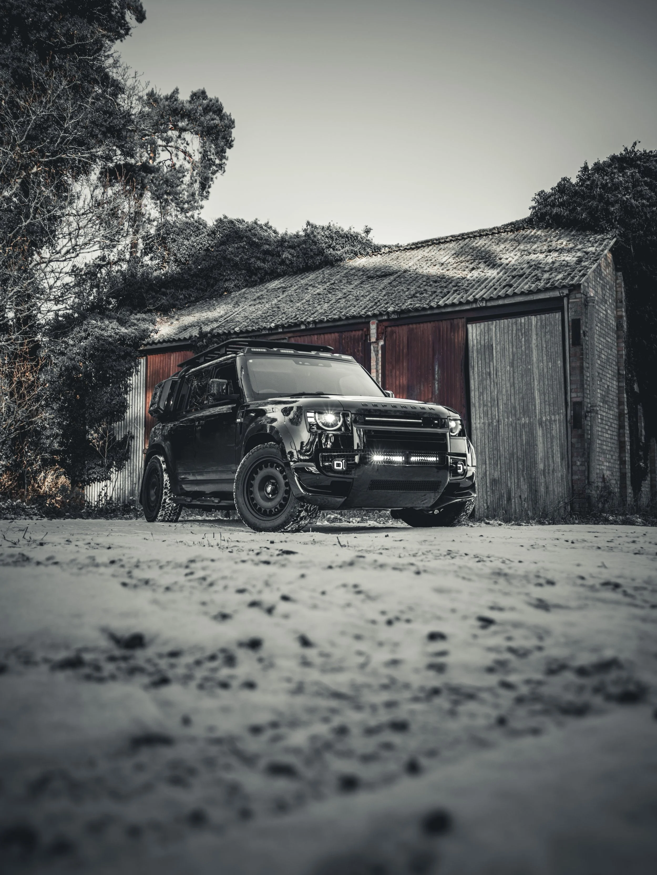 A black SUV parked in front of an old barn with a rusted metal door, surrounded by snow and leafless trees during winter.