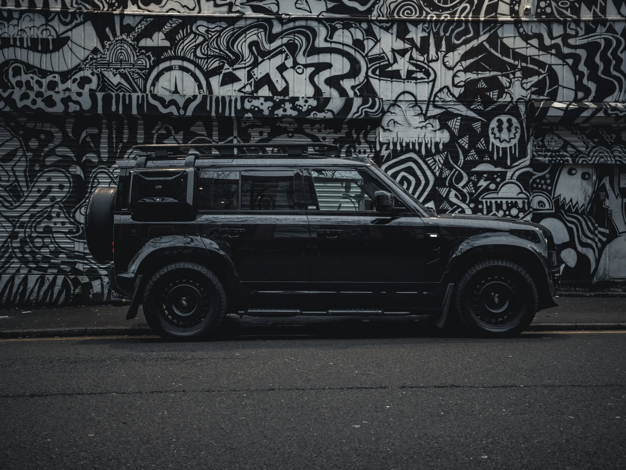 A black off-road vehicle parked in front of a black and white mural with abstract designs, skulls, and clock images.