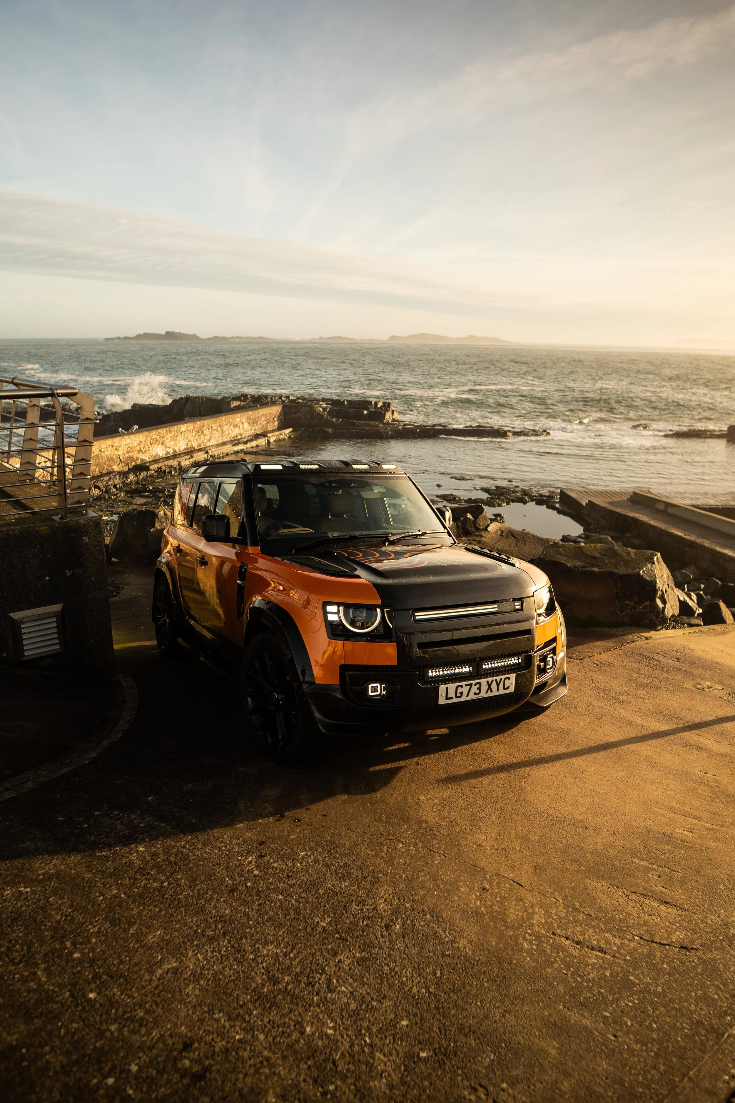 A black and orange Range Rover parked on a coastal road with rocks and the ocean in the background during sunset.