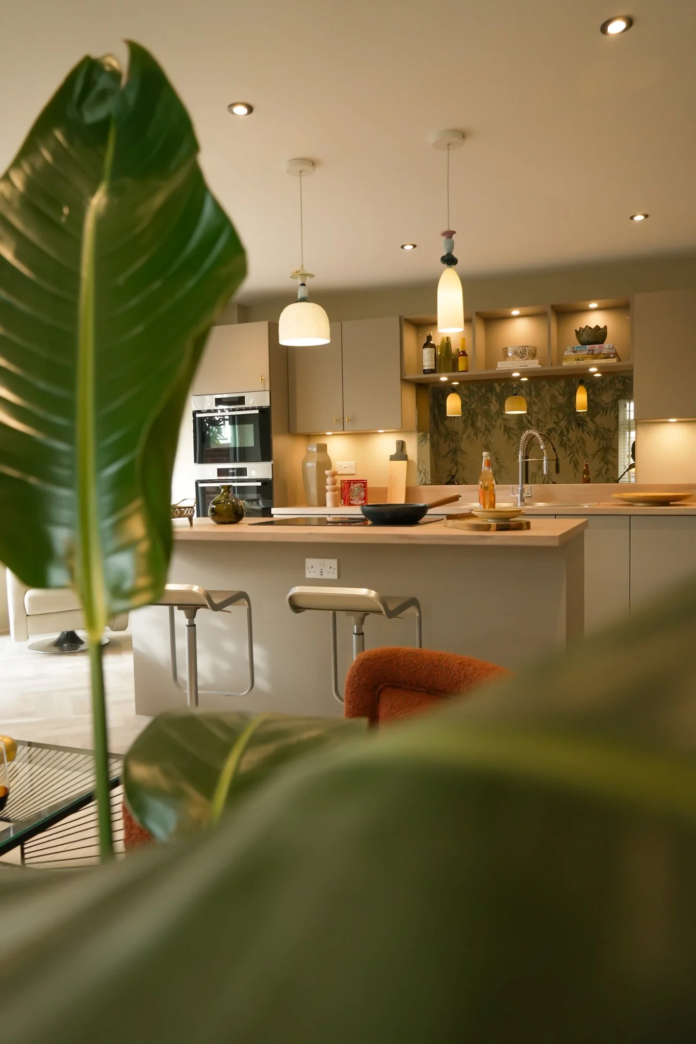A modern kitchen with pendant lighting, a kitchen island, and open shelves with decorative items. A large green leafy plant partially obscures the view.