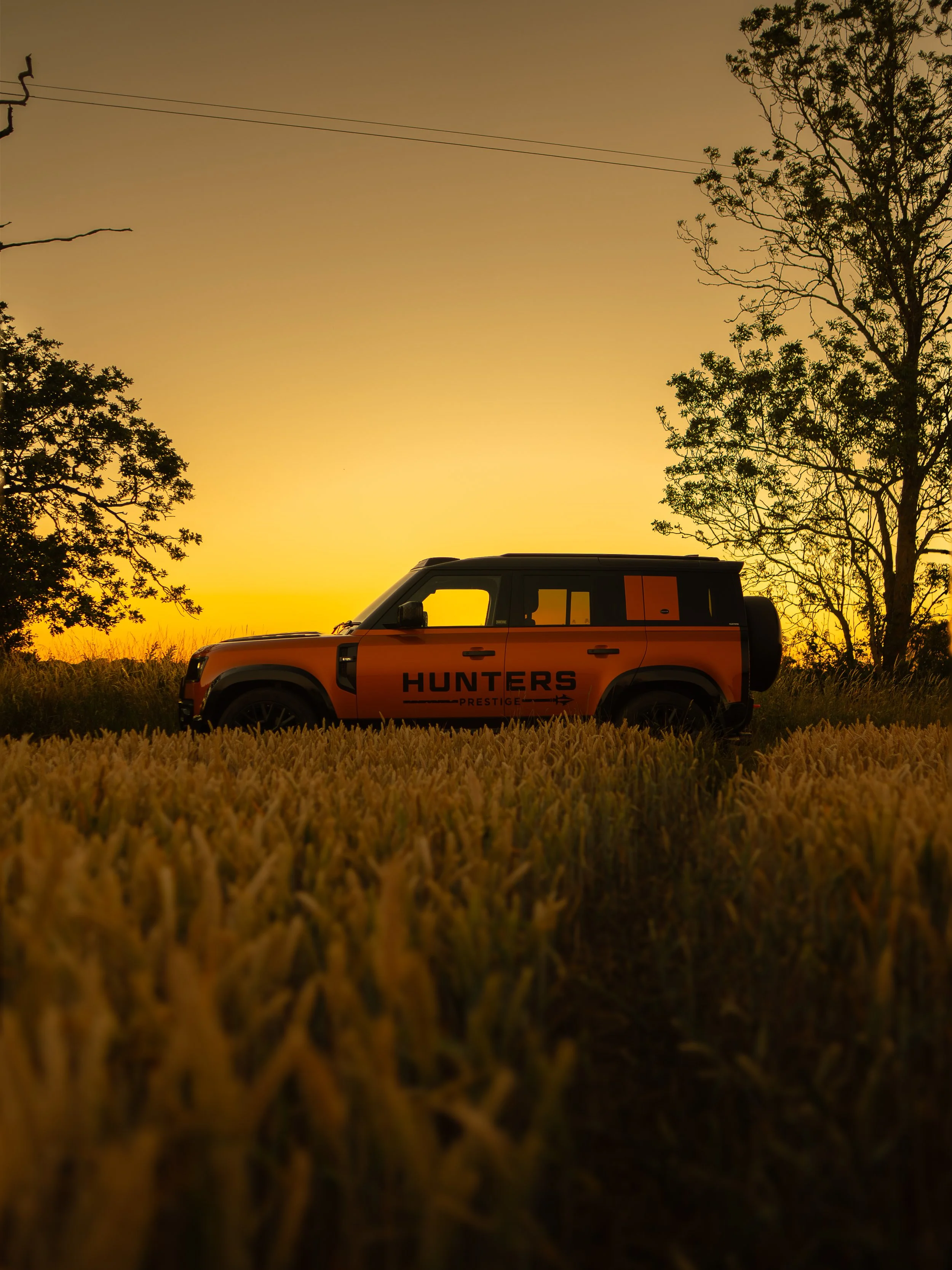A black and orange off-road vehicle with 'HUNTERS PRESTIGE' written on its side, parked on a grassy field during sunset with a tree on the right and power lines overhead.