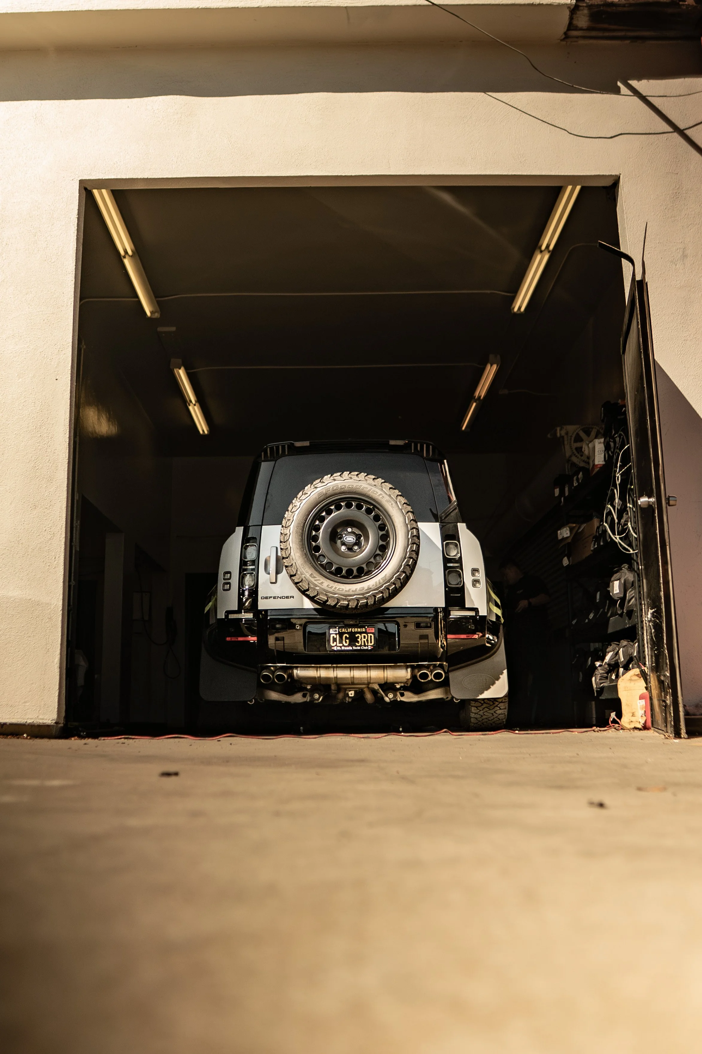 A black and white Land Rover Defender parked inside a garage with an open garage door, visible tire, and a California license plate.