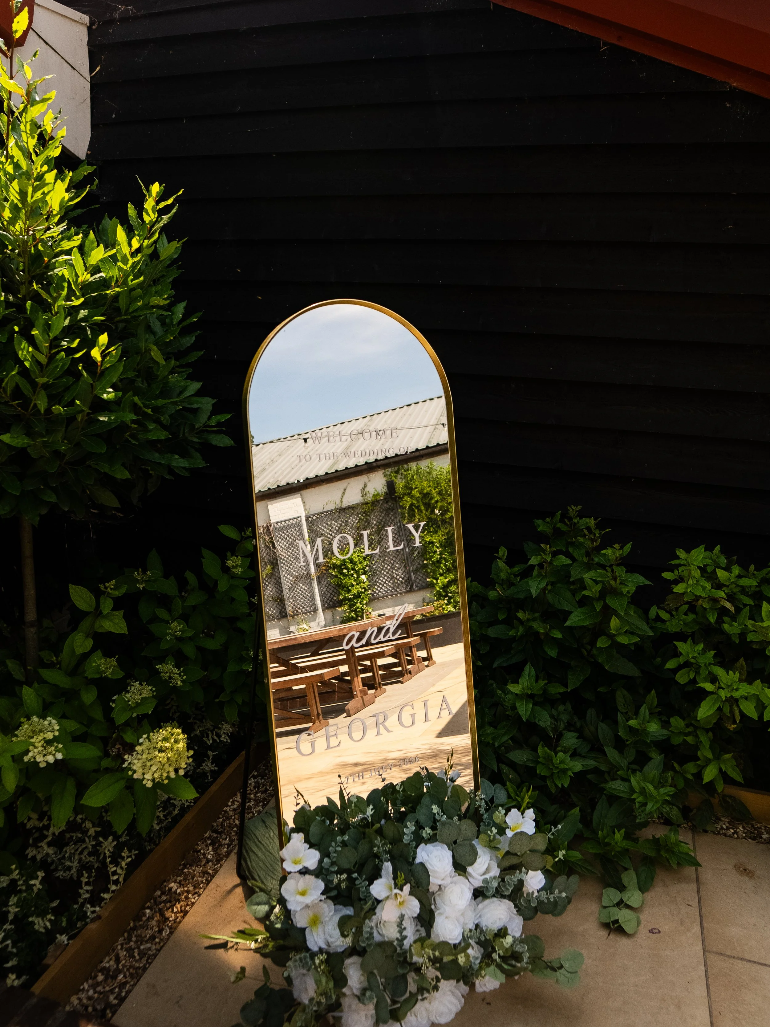 A decorative wedding sign with flowers at the base, surrounded by green bushes, with a black wooden wall and a part of a brown wooden roof in the background. The sign has a mirror with writing that says "Welcome to the wedding of Molly and Georgia, 17th July 2020."