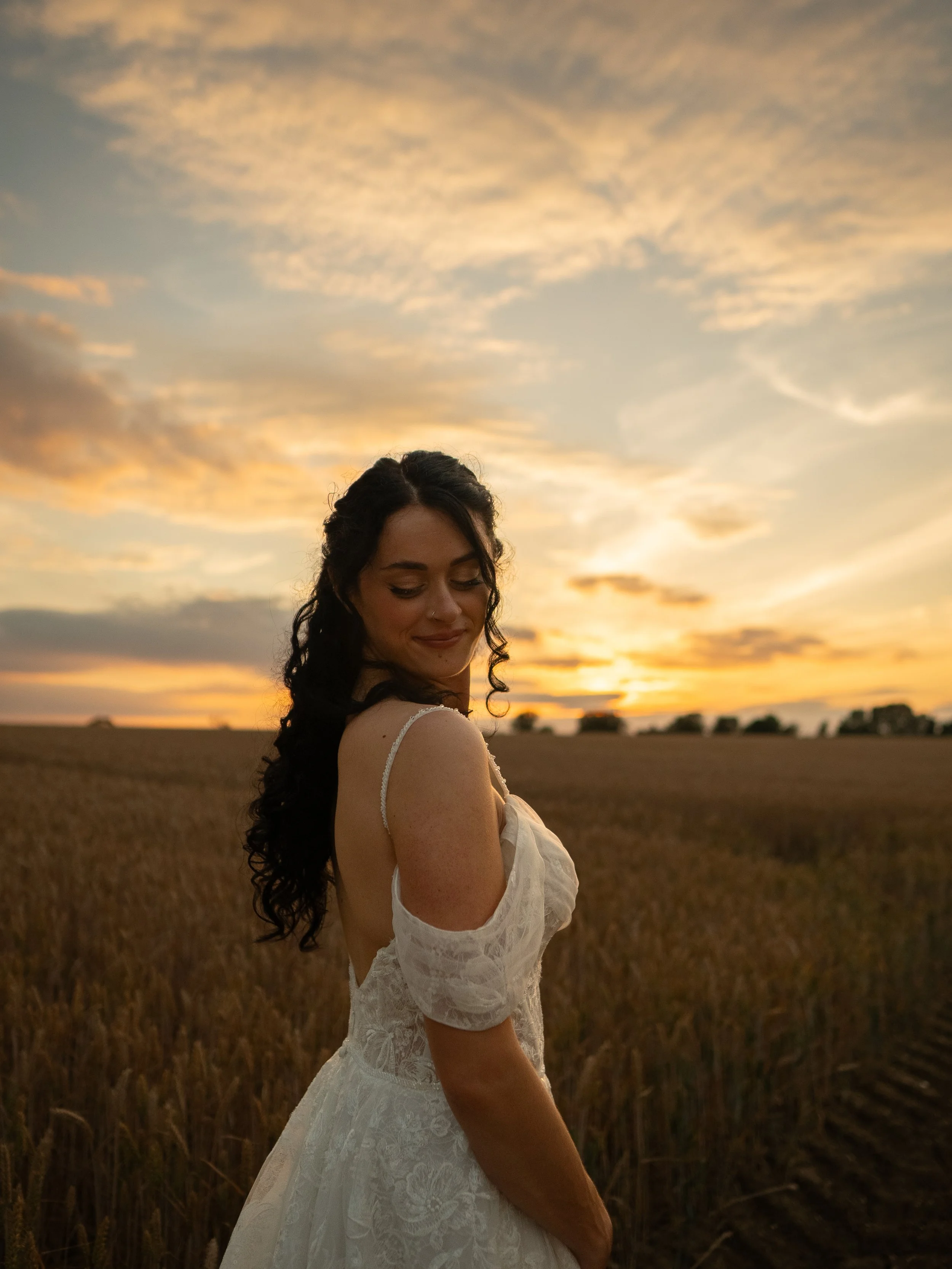 A young woman with dark, curly hair smiling with eyes closed, wearing a white lace dress, standing in a field during sunset with a sky filled with soft, orange clouds in the background.