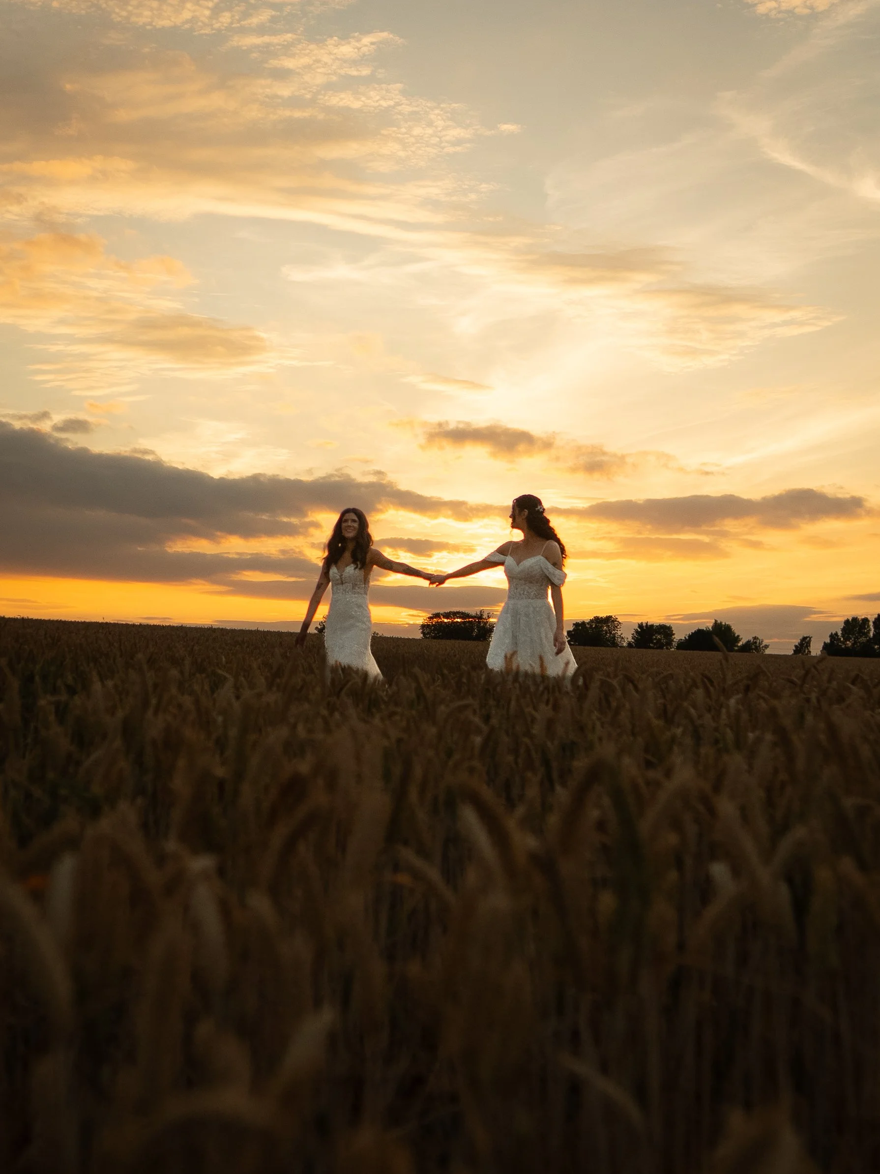 Two women in white dresses holding hands and smiling at sunset in a field.