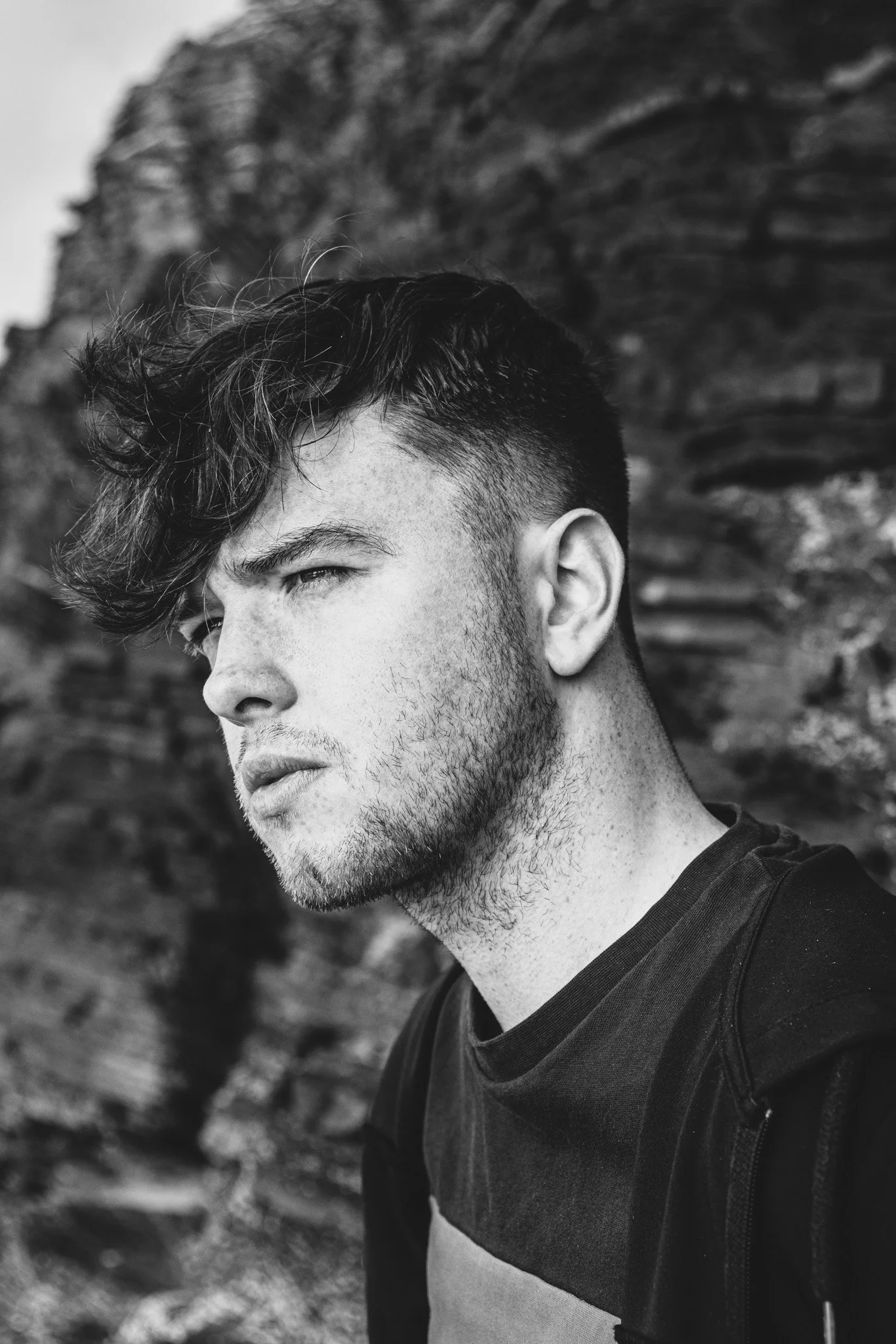 Black and white photo of a young man with short hair and a beard, looking to the side, outdoors with rocky background.