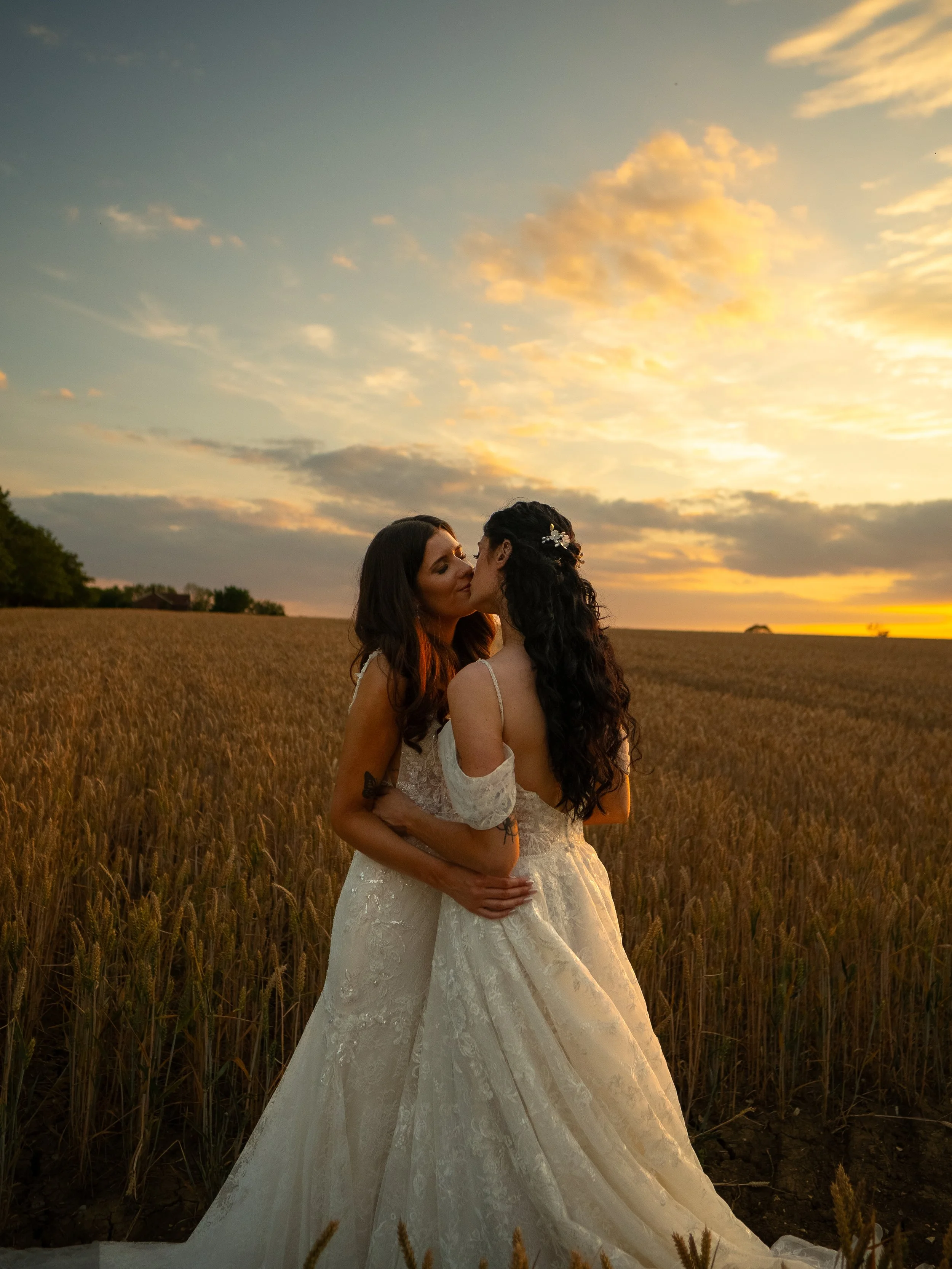 Two women in wedding dresses share a kiss and embrace in a wheat field during sunset.