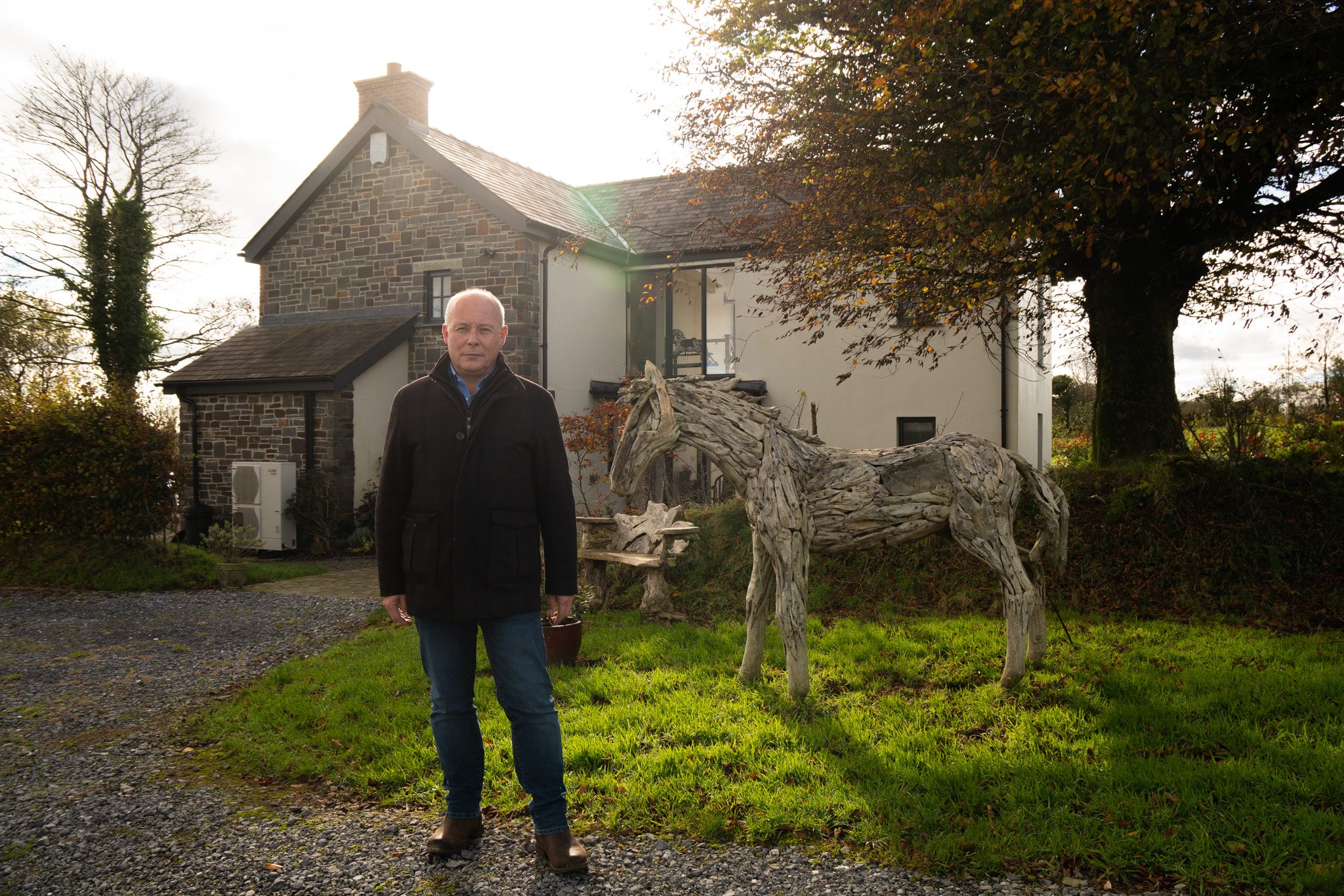 A man standing outside near a large tree and a wooden horse sculpture in front of a house with stone and white walls, during daytime with bright sunlight.