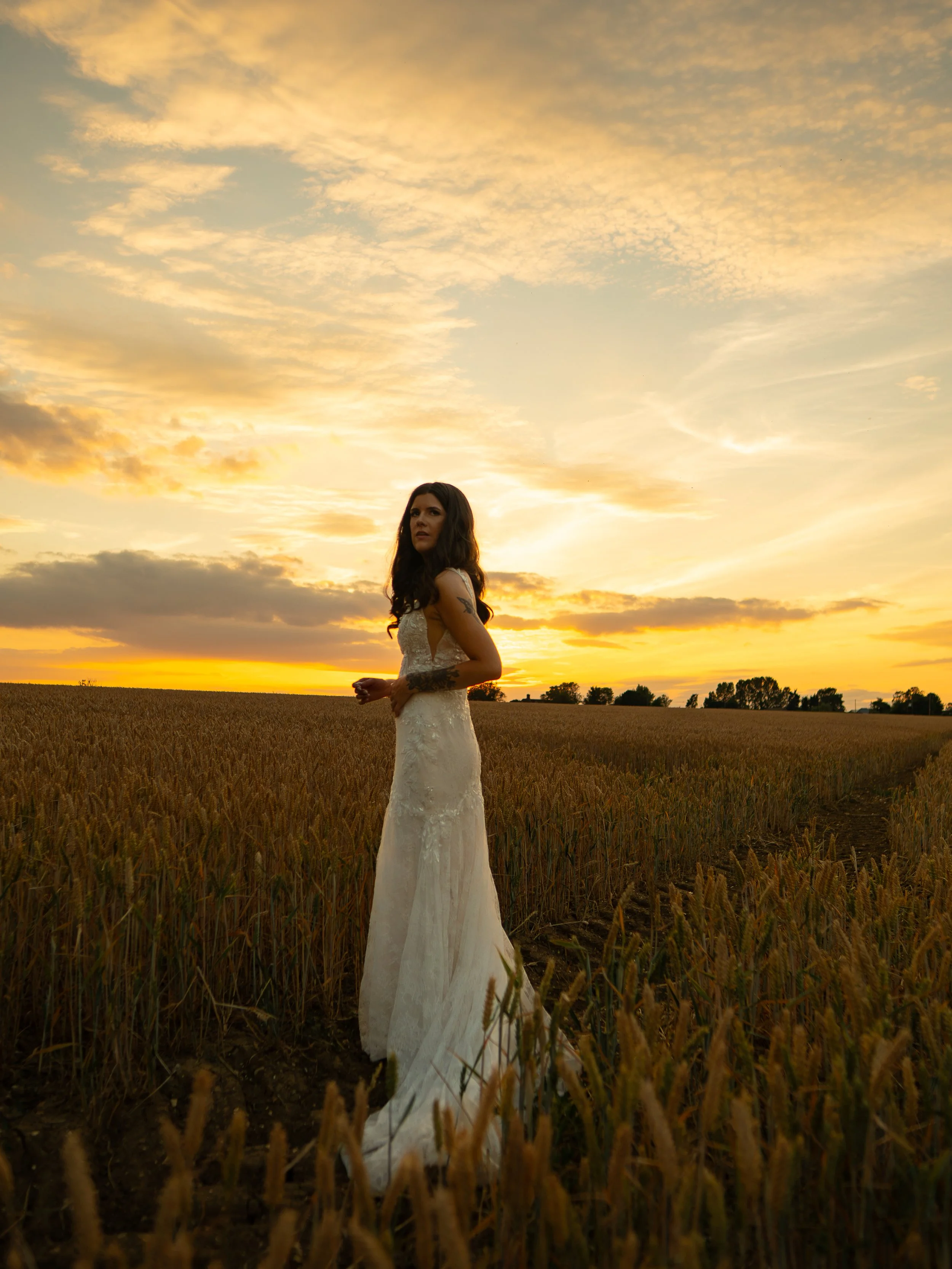 A woman in a white wedding dress standing in a wheat field at sunset with a sky filled with clouds.