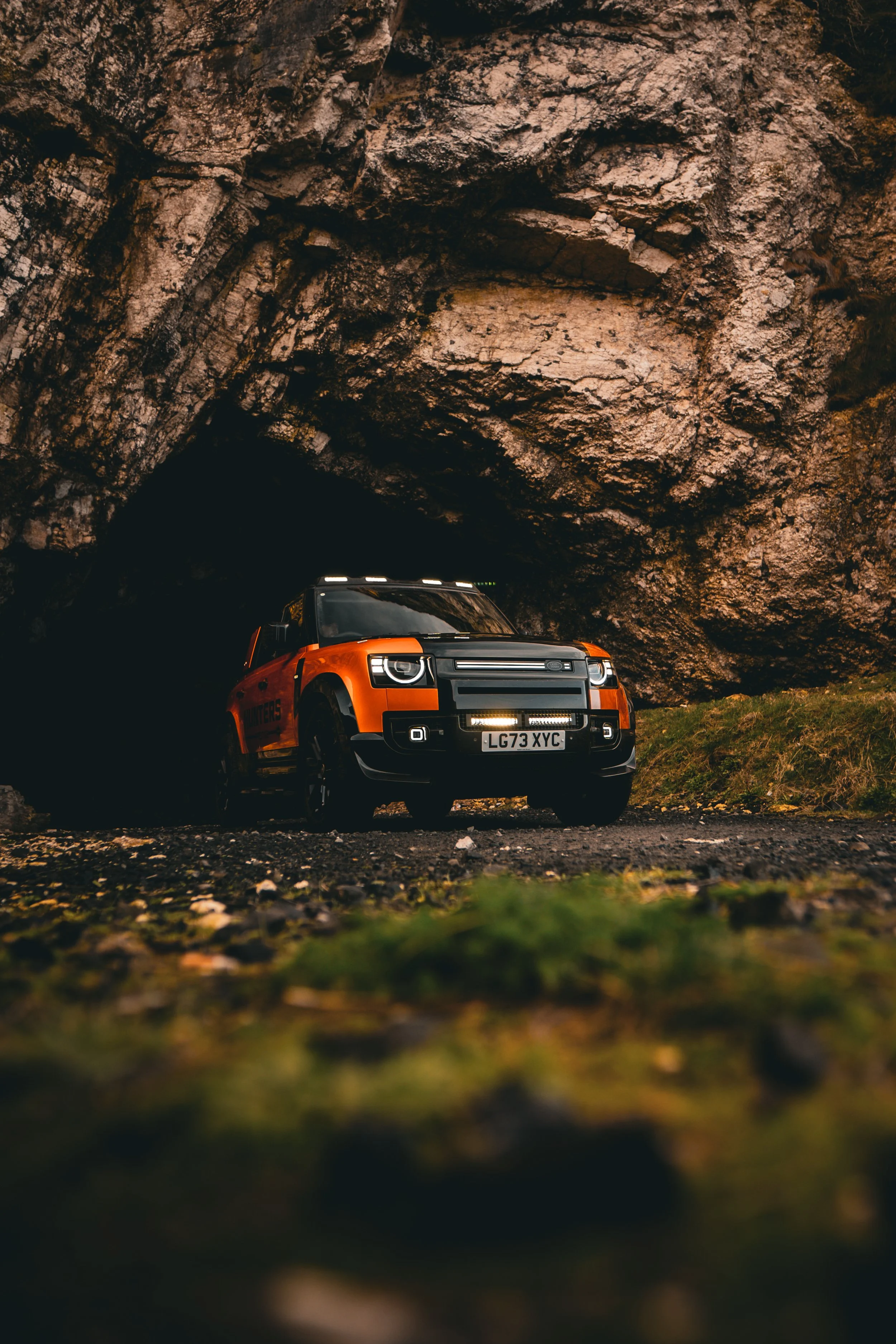 A bright orange off-road vehicle parked at the entrance of a dark cave, surrounded by a rocky terrain and greenery.